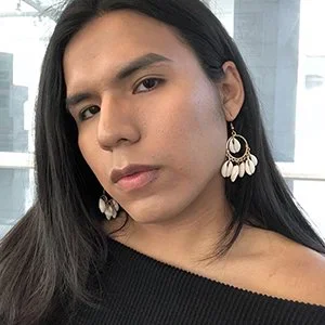 Jimena Lucero, a queer brown Latinx woman with her hair parted down the middle; she is wearing large dangly earrings, a black off the shoulder top, and has her face slightly turned away from the camera.