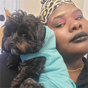 Rise, a Black disabled femme, is pictured from the chest up in a black hoodie and a leopard print headwrap. Jelly, a black cockapoo, is sitting on their forearm in a teal hoodie. They are both looking forward into the camera.