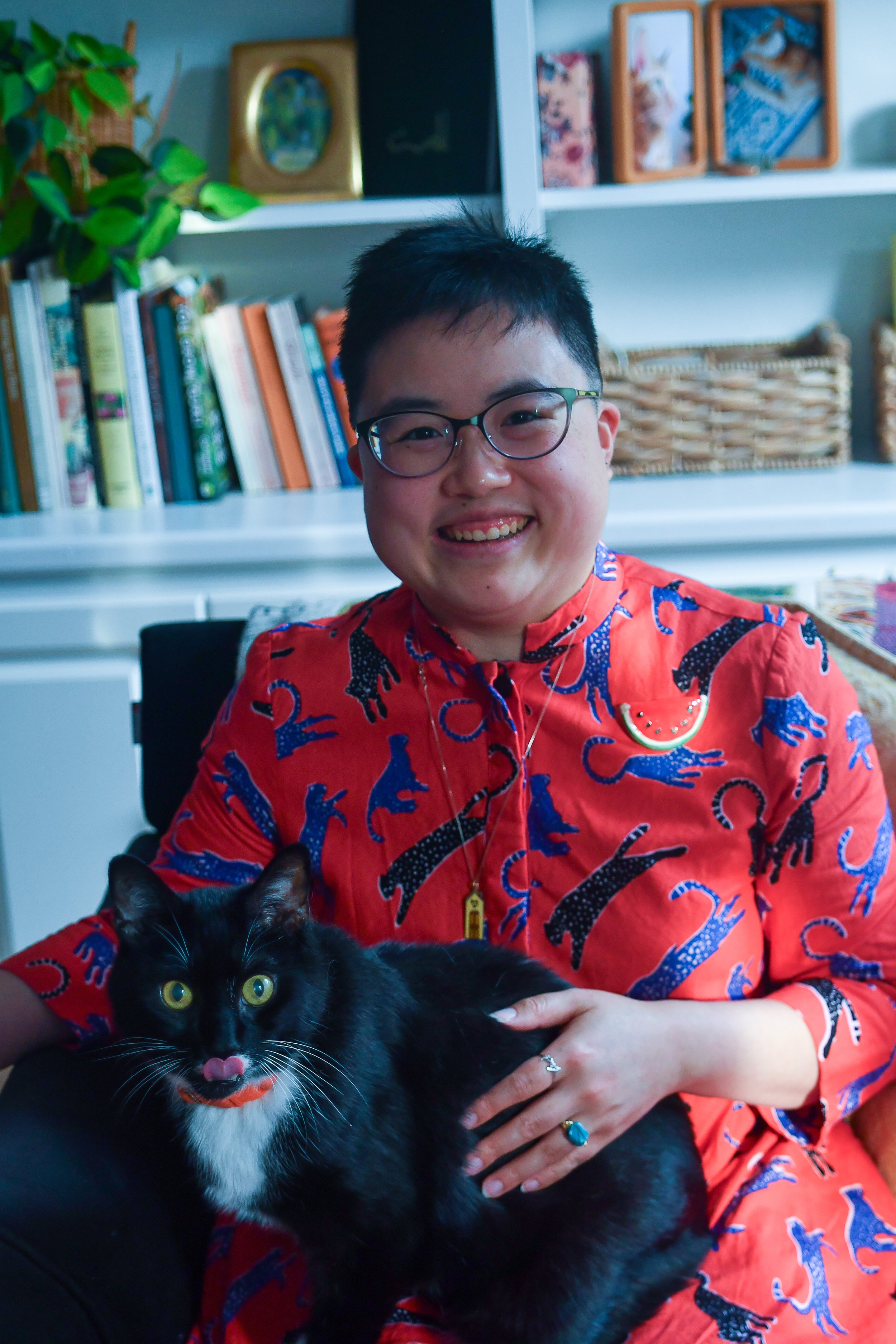 Ly Xīnzhèn, an East Asian person in their thirties with short black hair and glasses, is smiling from an armchair. They have a tuxedo cat on their lap with his tongue sticking out and are resting a hand with a turquoise ring on the cat's side.