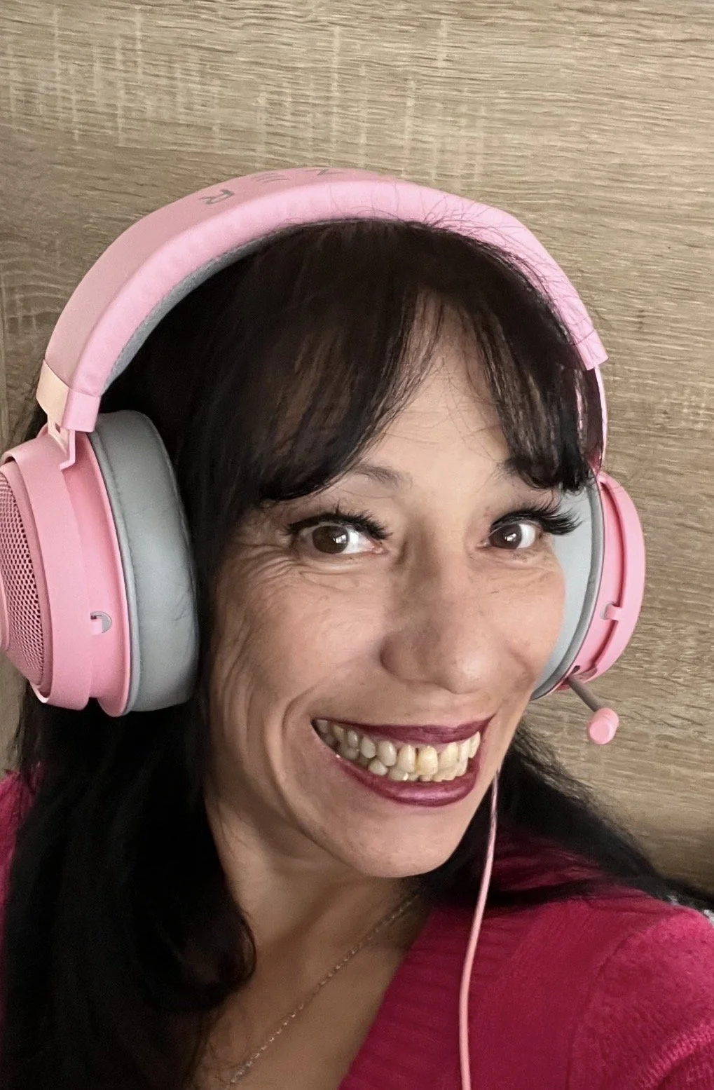 A Latina woman with fair skin, brown hair, and brown eyes smiles at the camera. She is wearing pink headphones and a dark pink sweater.