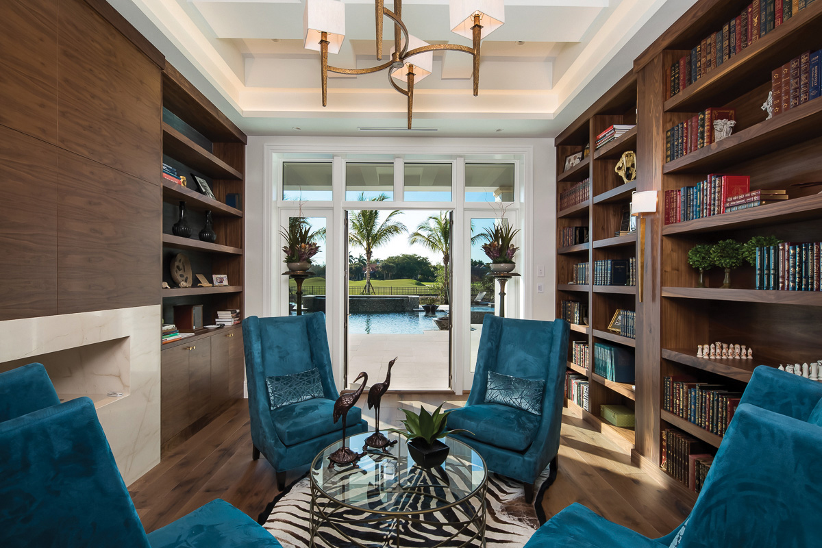  The home’s library features modern, dark wood built-in shelves and cabinets which are juxtaposed by the bright colors of the four accent chairs and fun lighting choices. Here, the other side of the see-through fireplace is visible and has been clad 