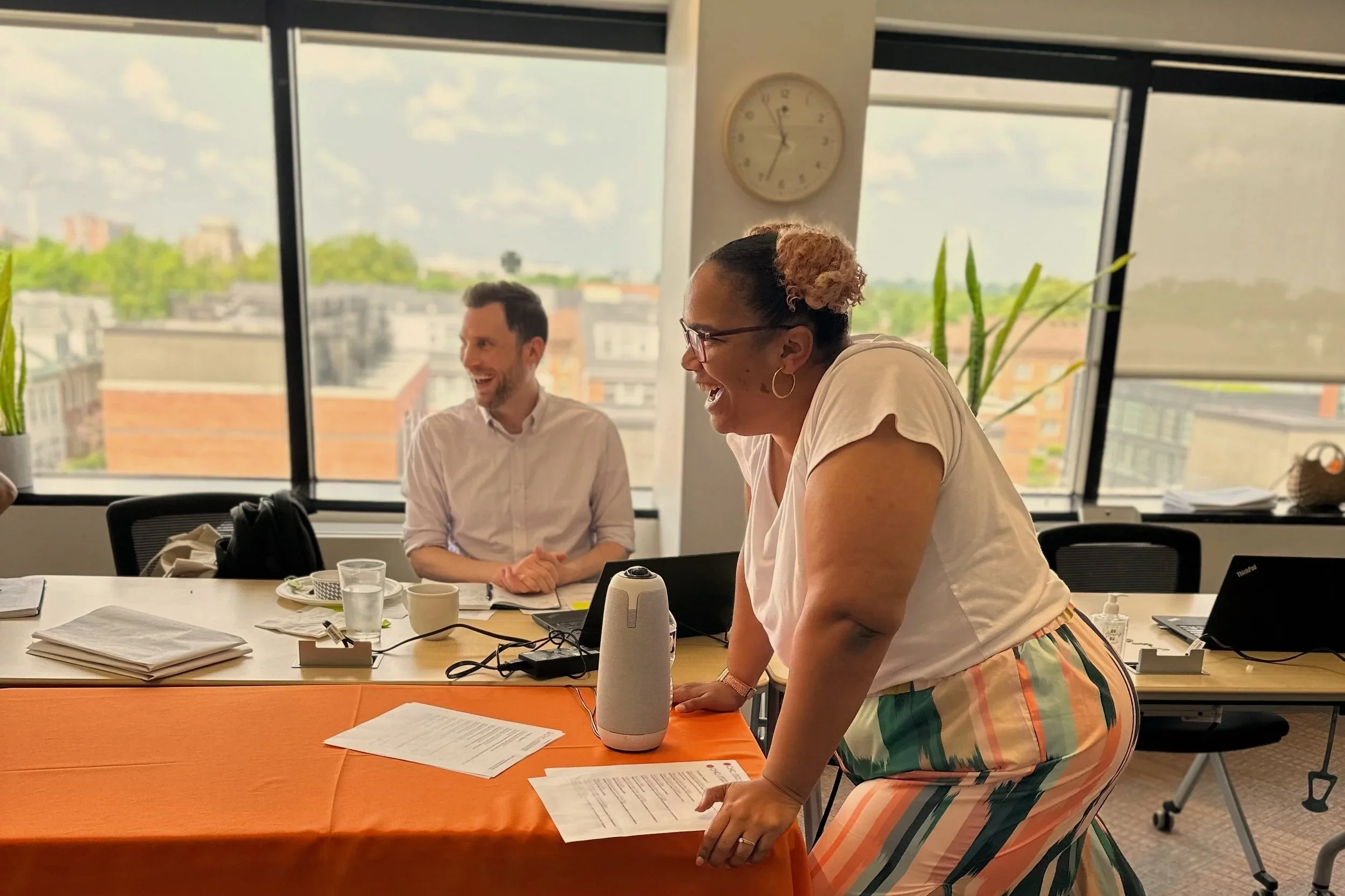 People in a meeting room laughing, with two women and a man, with papers, laptops, drinks, and a speaker on the table, large windows with a city view, and a clock showing 11:50.