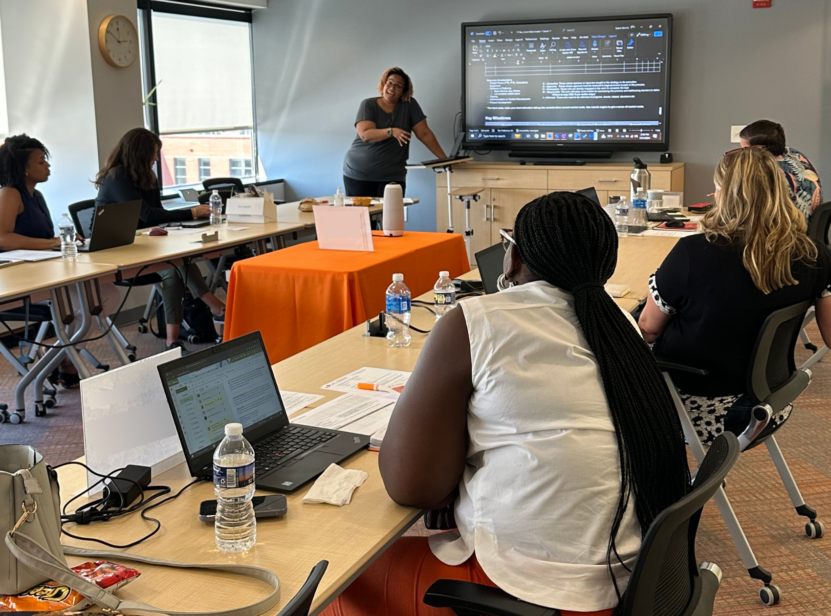 A woman is standing and presenting in a conference room with a large screen, while several people are seated at tables with laptops and water bottles, listening to her.