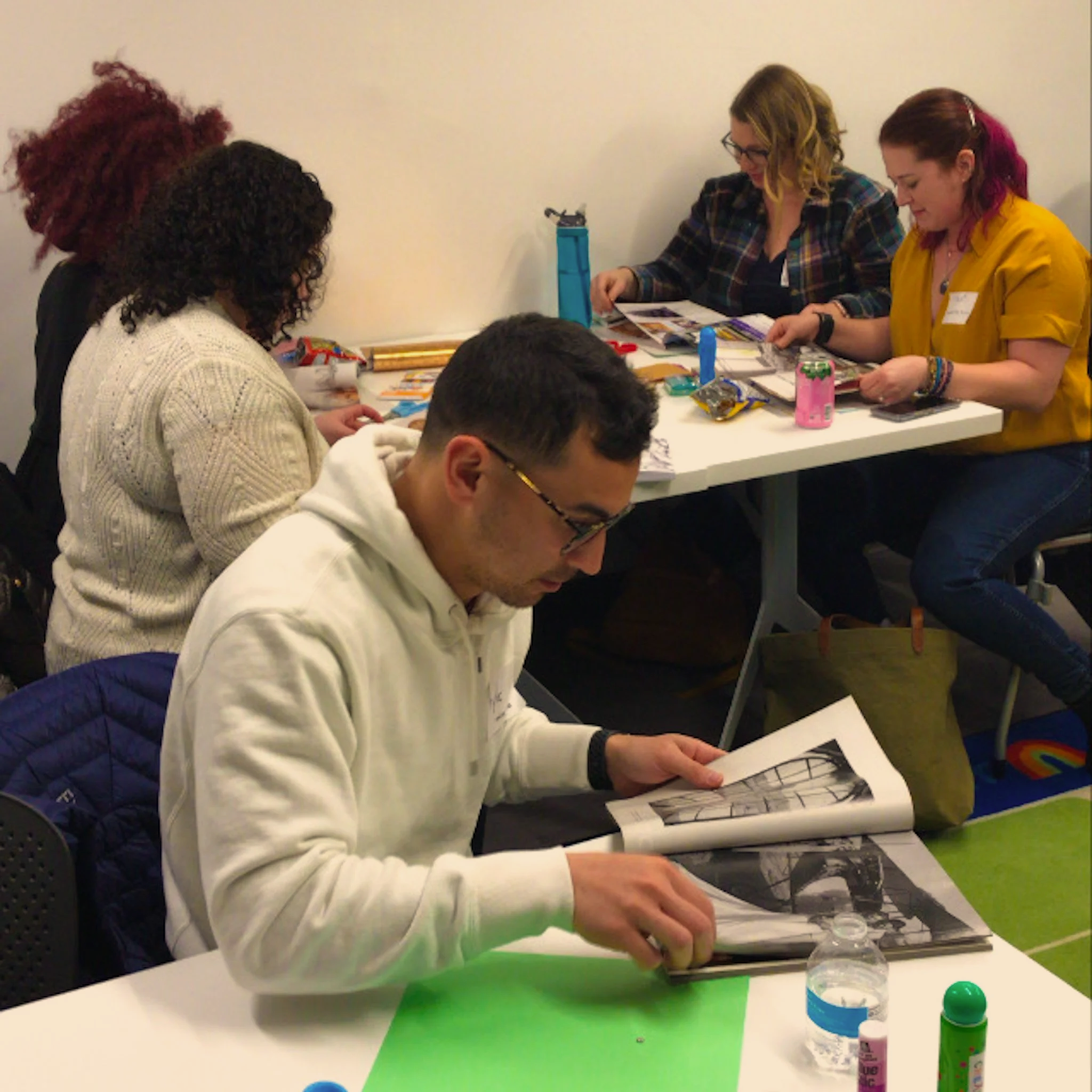 People sitting at a table engaging in an activity with craft supplies, including papers, bottles, and other materials, in an indoor setting.