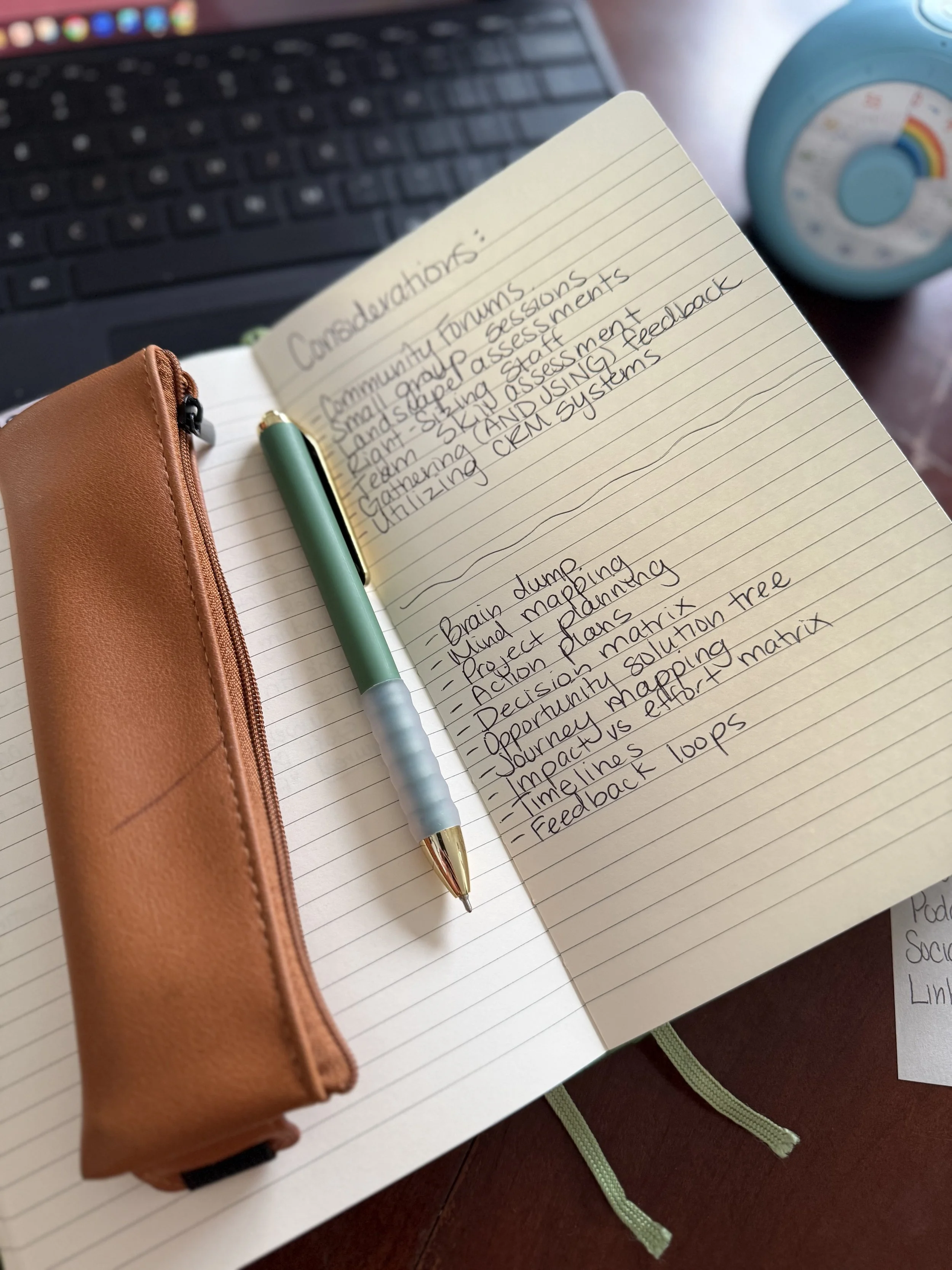 Open notebook with handwritten notes on considerations for community forums, including topics like brain dump, project plans, and feedback loops. A green pen rests on the page, and a leather zipper case is nearby. In the background, a black laptop keyboard and a blue clock with a rainbow design are visible.