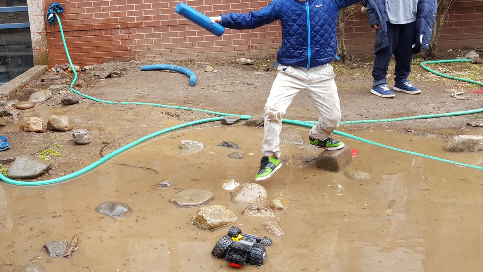  A child steps gingerly from one rock to another, maneuvering above a flooded muddy area. Another child stands on not flooded ground nearby, and a green hose snakes its way around the background of the image, and a toy truck sits partly submerged in 