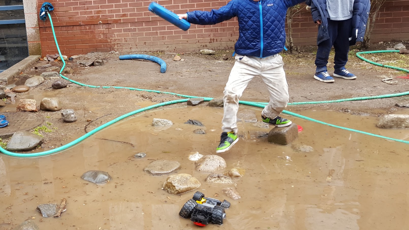 A child steps gingerly from one rock to another, maneuvering above a flooded muddy area. Another child stands on not flooded ground nearby, and a green hose snakes its way around the background of the image, and a toy truck sits partly submerged in the puddle in the foreground.