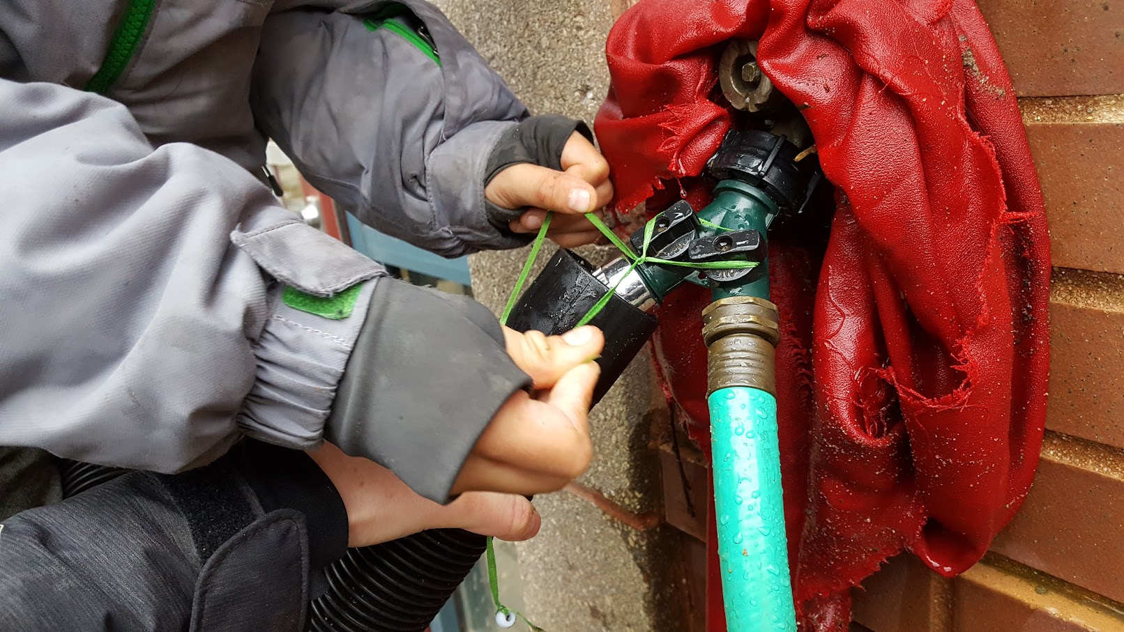  Closeup of a hose attached to a faucet on a brick and concrete wall. a child’s hands maneuver some string around the knobs of the faucet. 