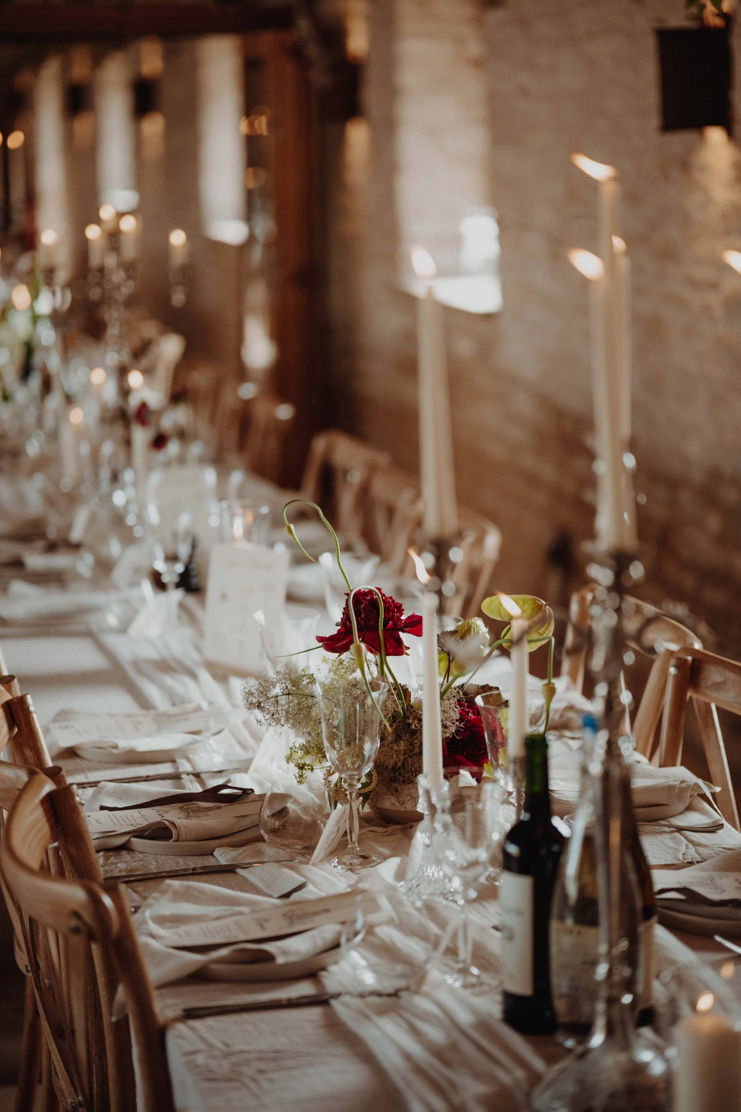 A decorated dining table with floral centerpieces, tall candles, wine bottles, and place settings in a rustic indoor setting.