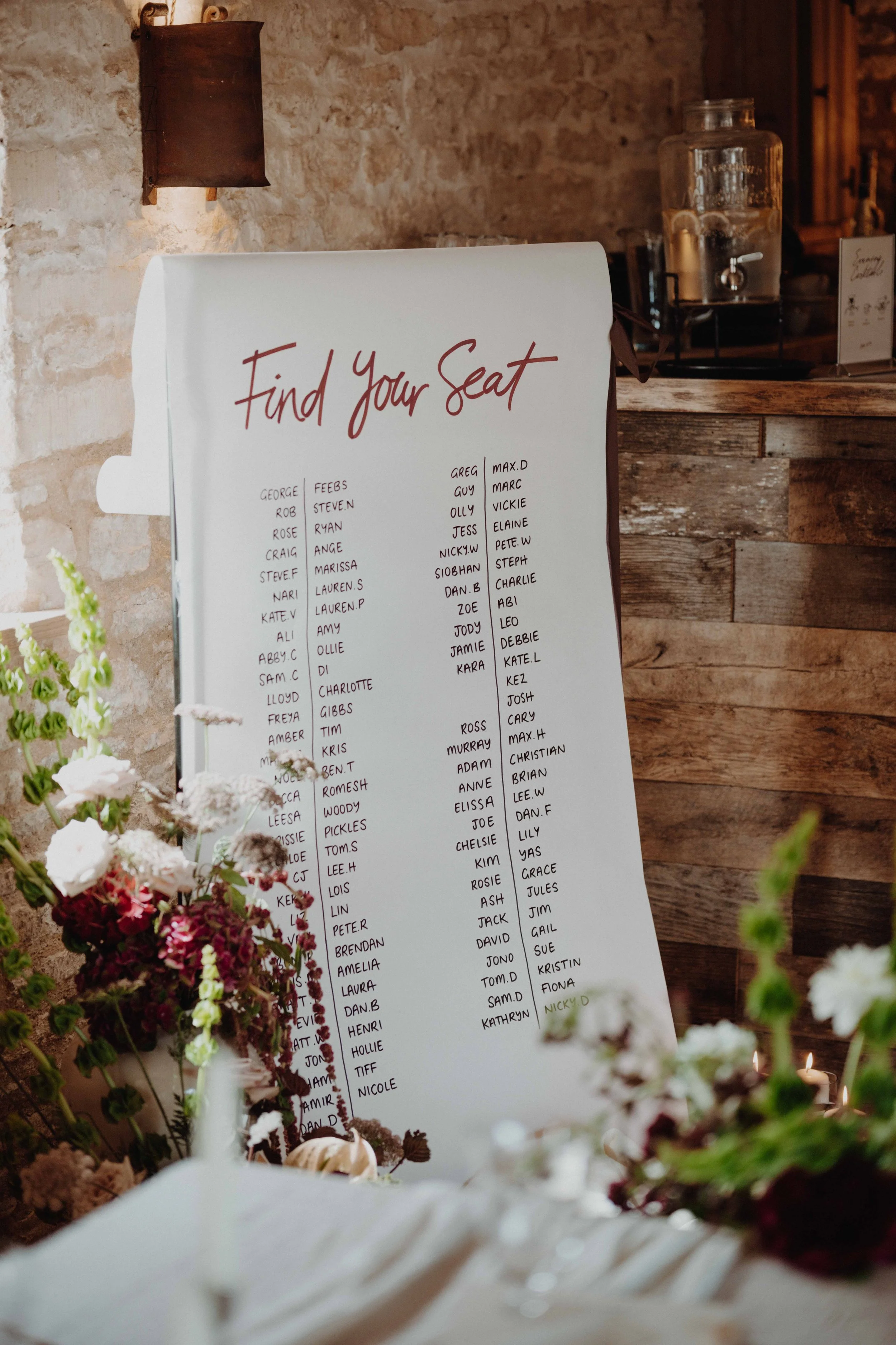 A large white sign with red cursive text that says 'Find Your Seat' and a list of guest names organized in columns, with some names highlighted in green. The sign is set against a rustic brick wall with floral decorations in the foreground and a wate