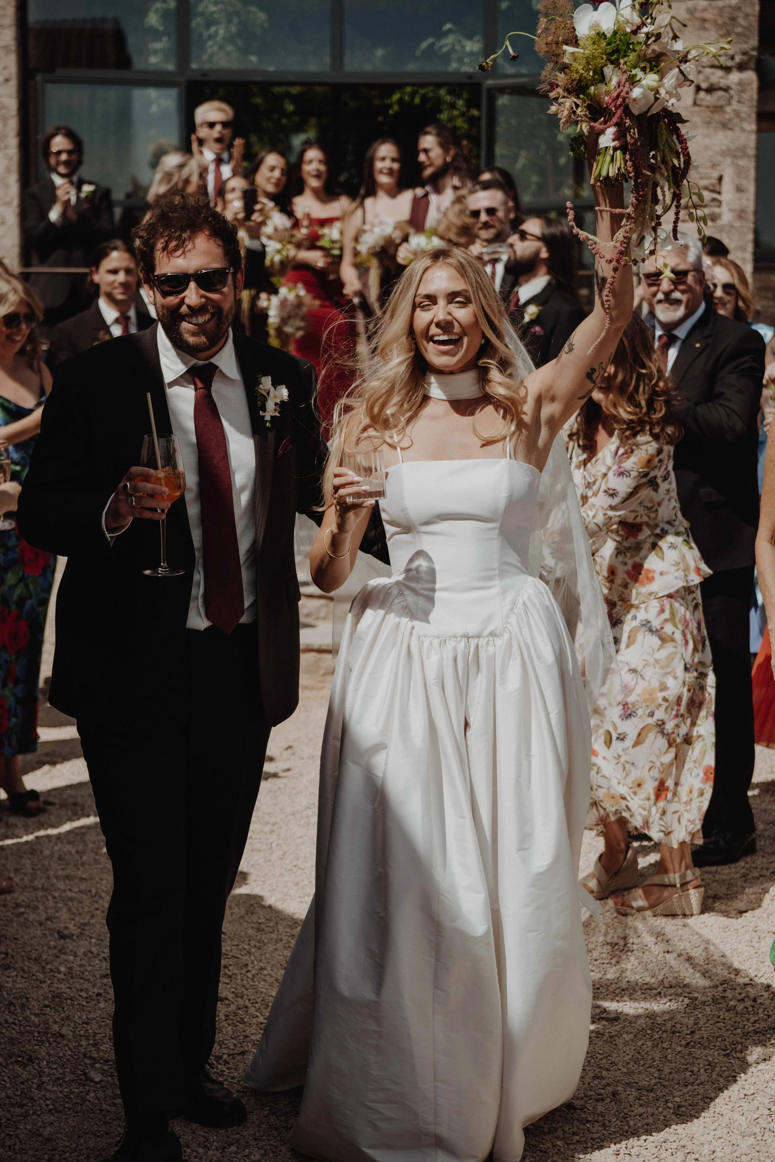 A bride in a white wedding dress and a groom in a black suit celebrating outside with fireworks. Guests cheer and smile around them, some holding drinks, in front of a building with large glass windows.