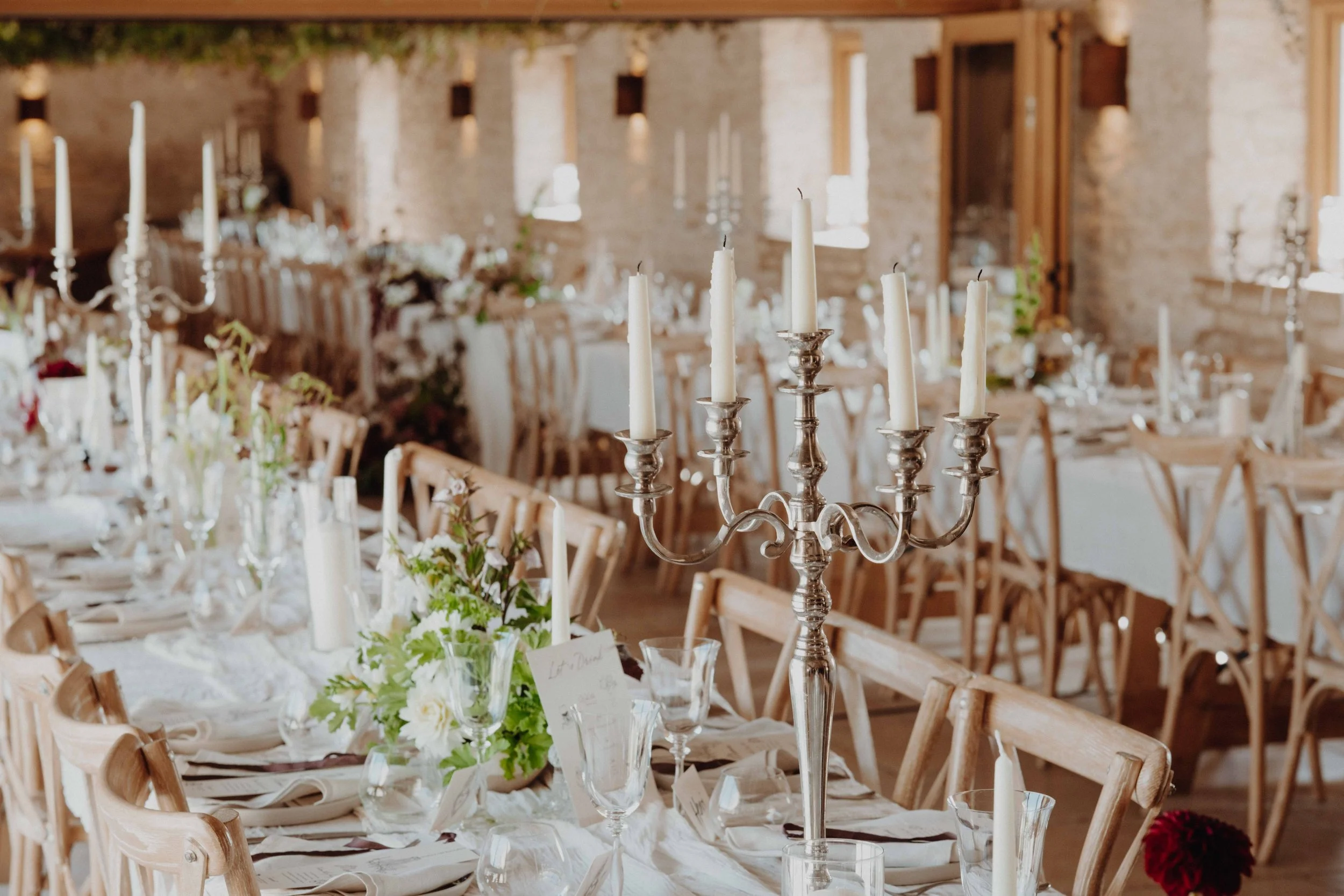 A wedding reception setup with wooden chairs around tables decorated with white tablecloths, floral centerpieces, and tall silver candelabras with unlit white candles inside a rustic venue with exposed brick walls and natural lighting.