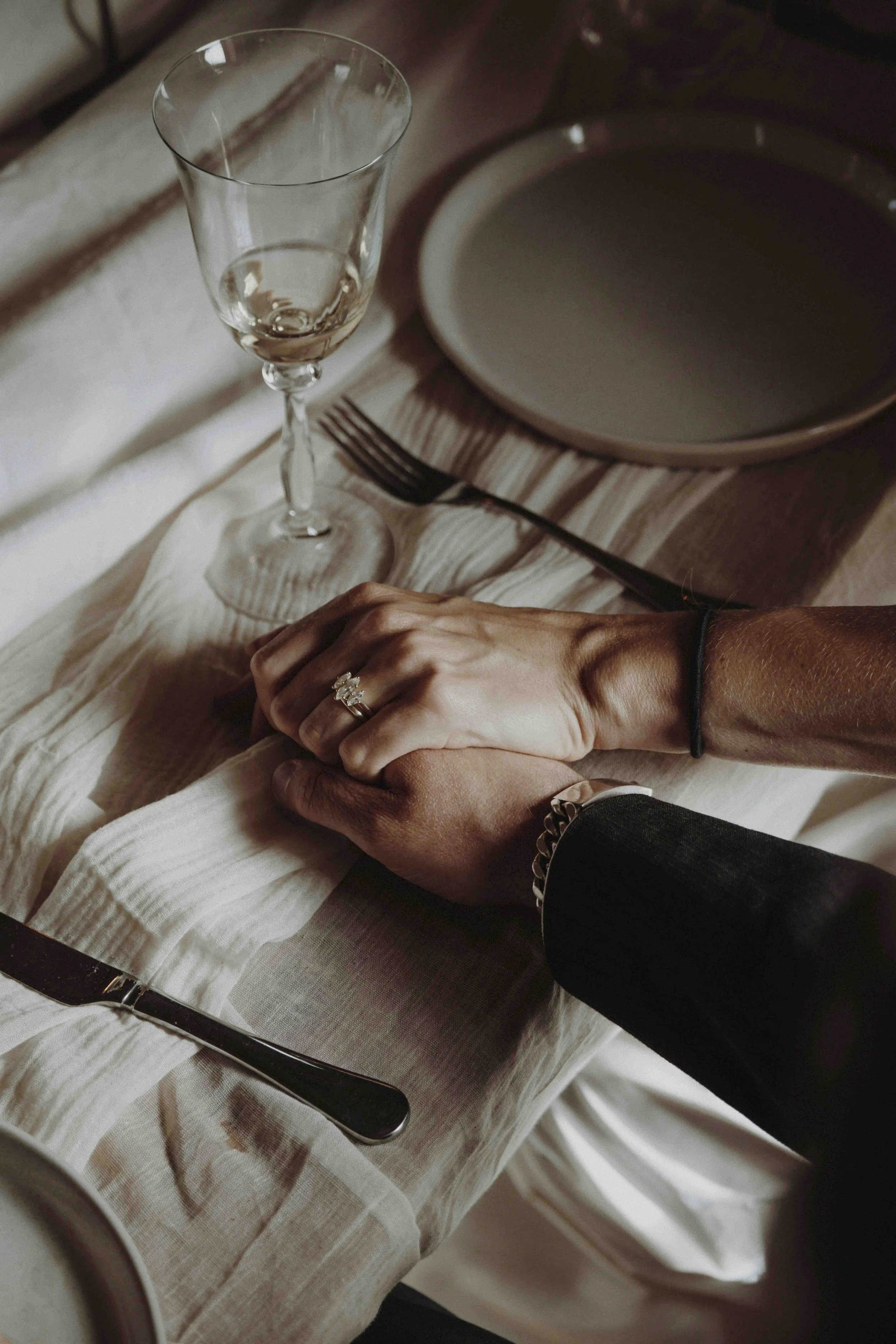 Holding hands at a formal dinner table with a wedding ring, a glass of wine, and place settings.