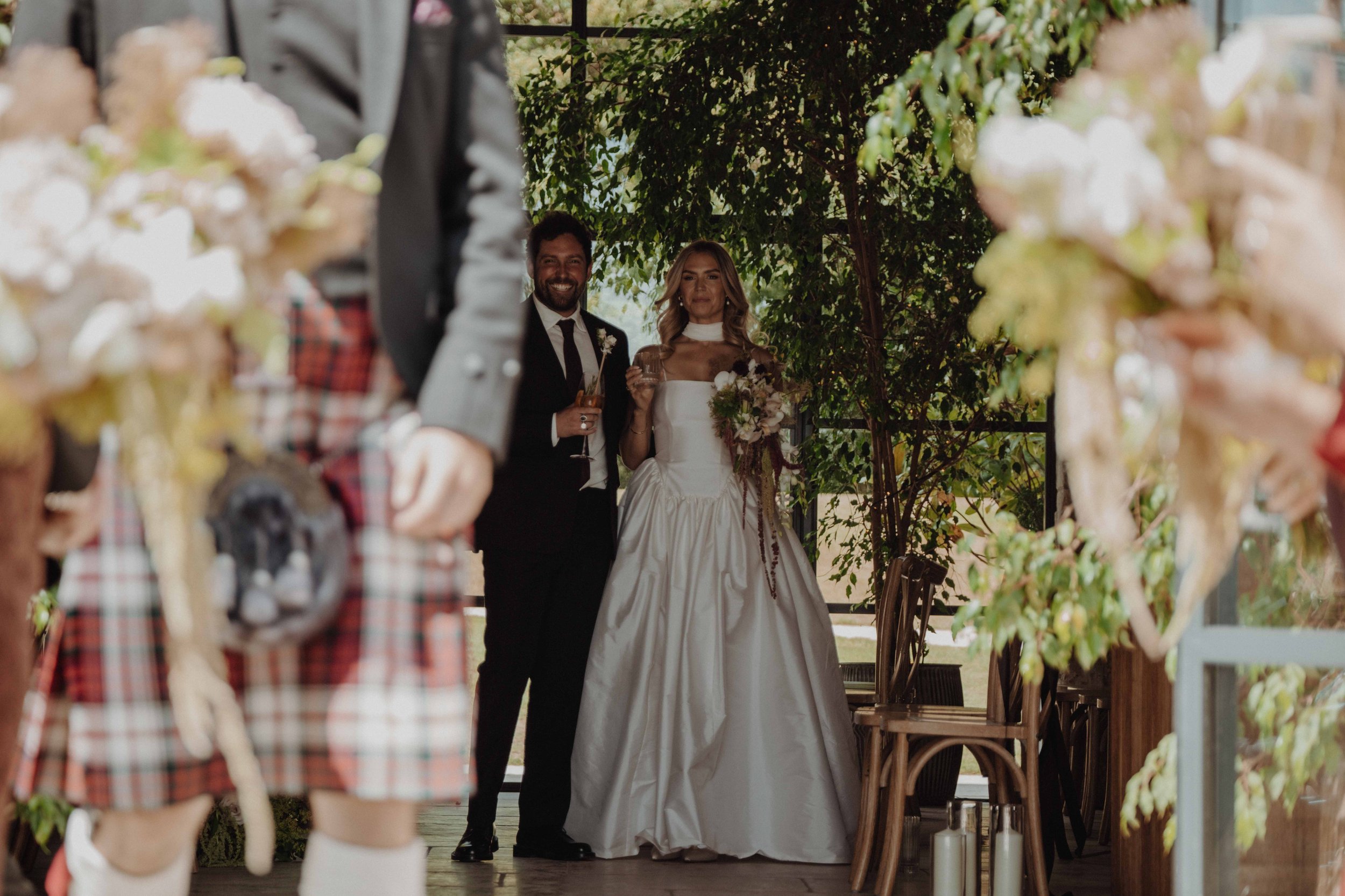 Bride and groom standing together, holding glasses, at their wedding reception with guests and floral decorations nearby.