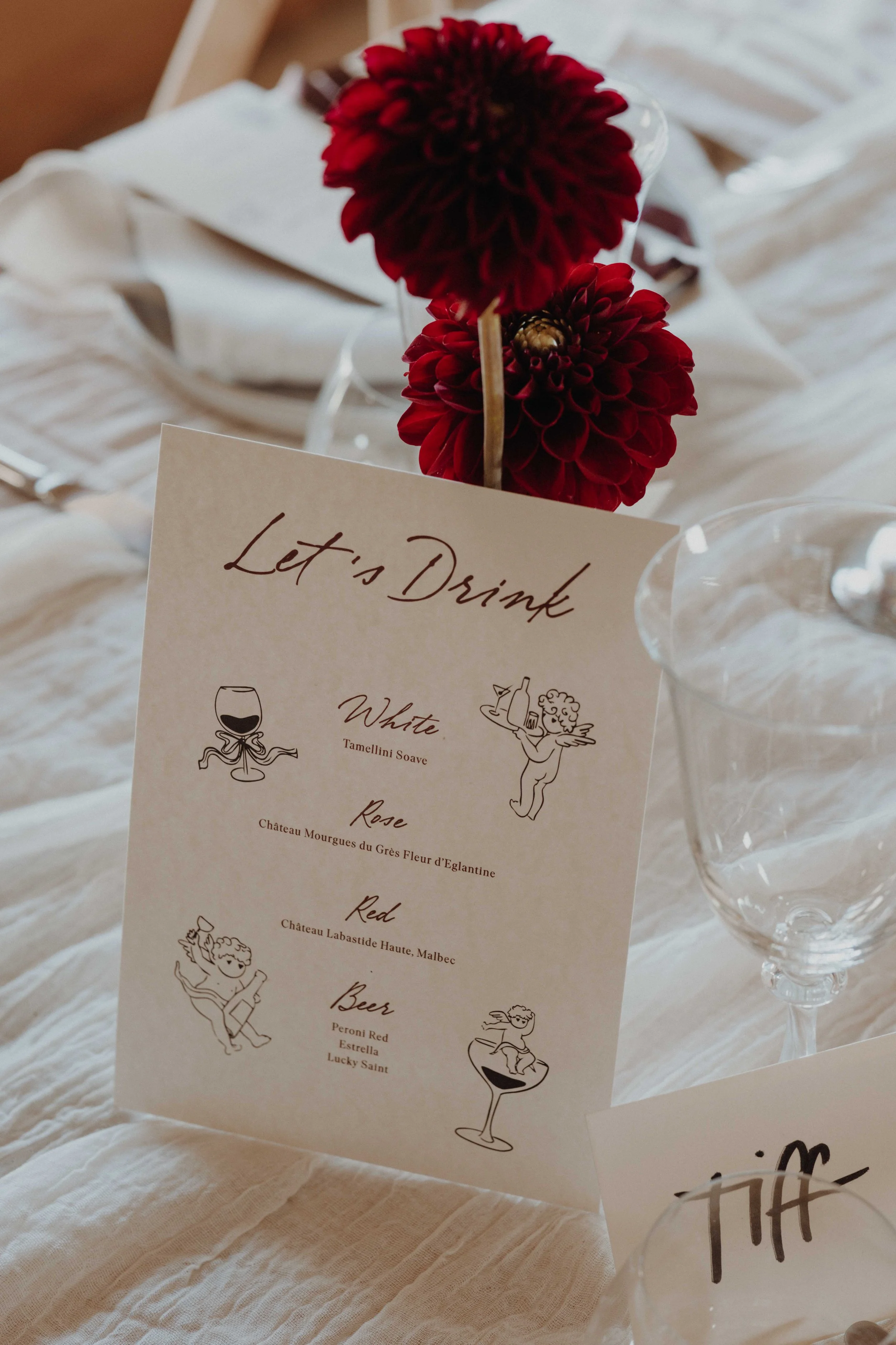 A drink menu card titled 'Let's Drink' with illustrations of different types of beverages and their names, placed on a table decorated with red flowers in a vase and a glass of water.