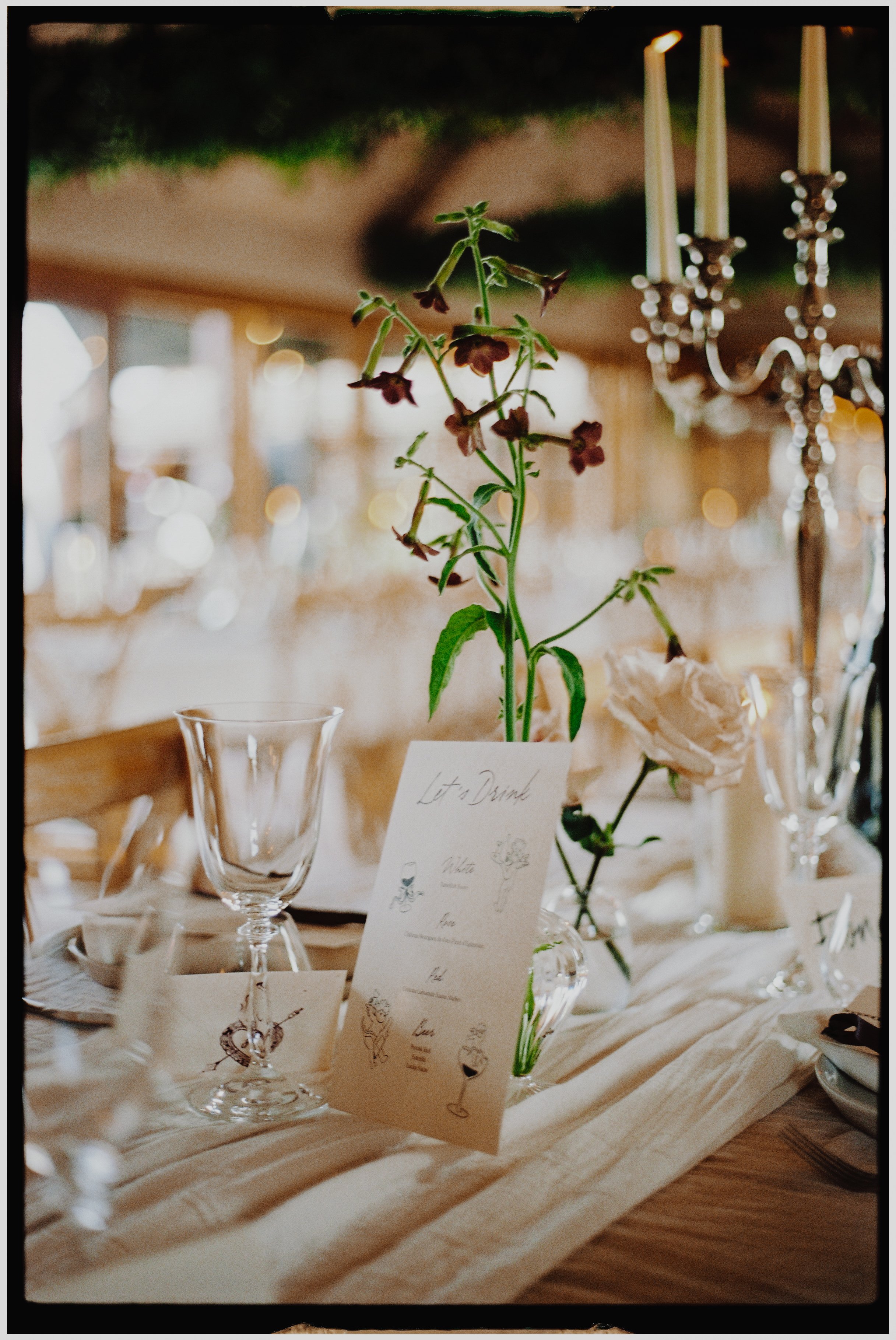 Close-up of a table setting with a tall flower arrangement, wine glass, and cocktail menu, in a warmly lit venue with hanging candles and blurred background.