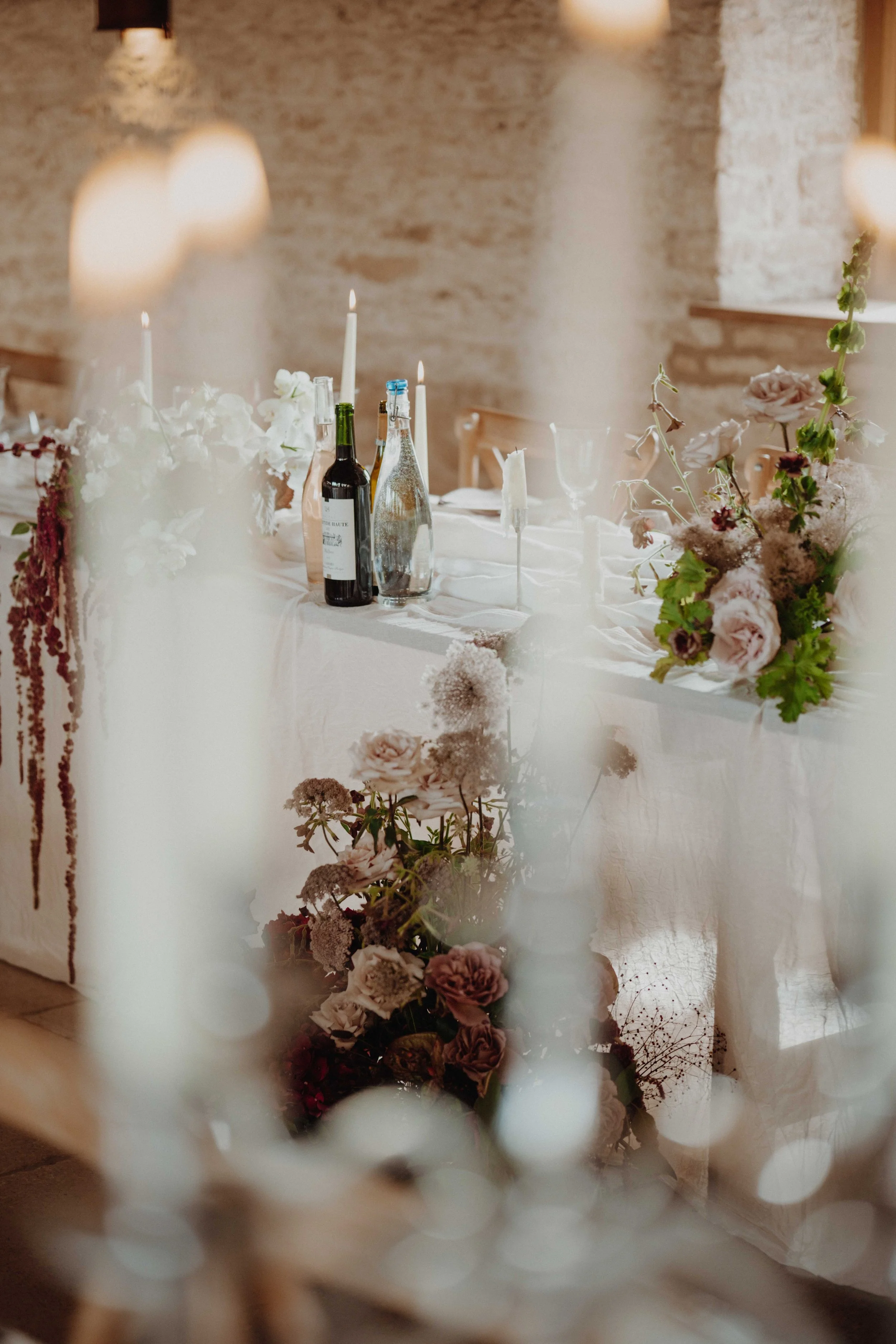 A decorated table set for a special occasion with floral arrangements, candles, wine bottles, and glasses in a rustic room with a brick wall background.