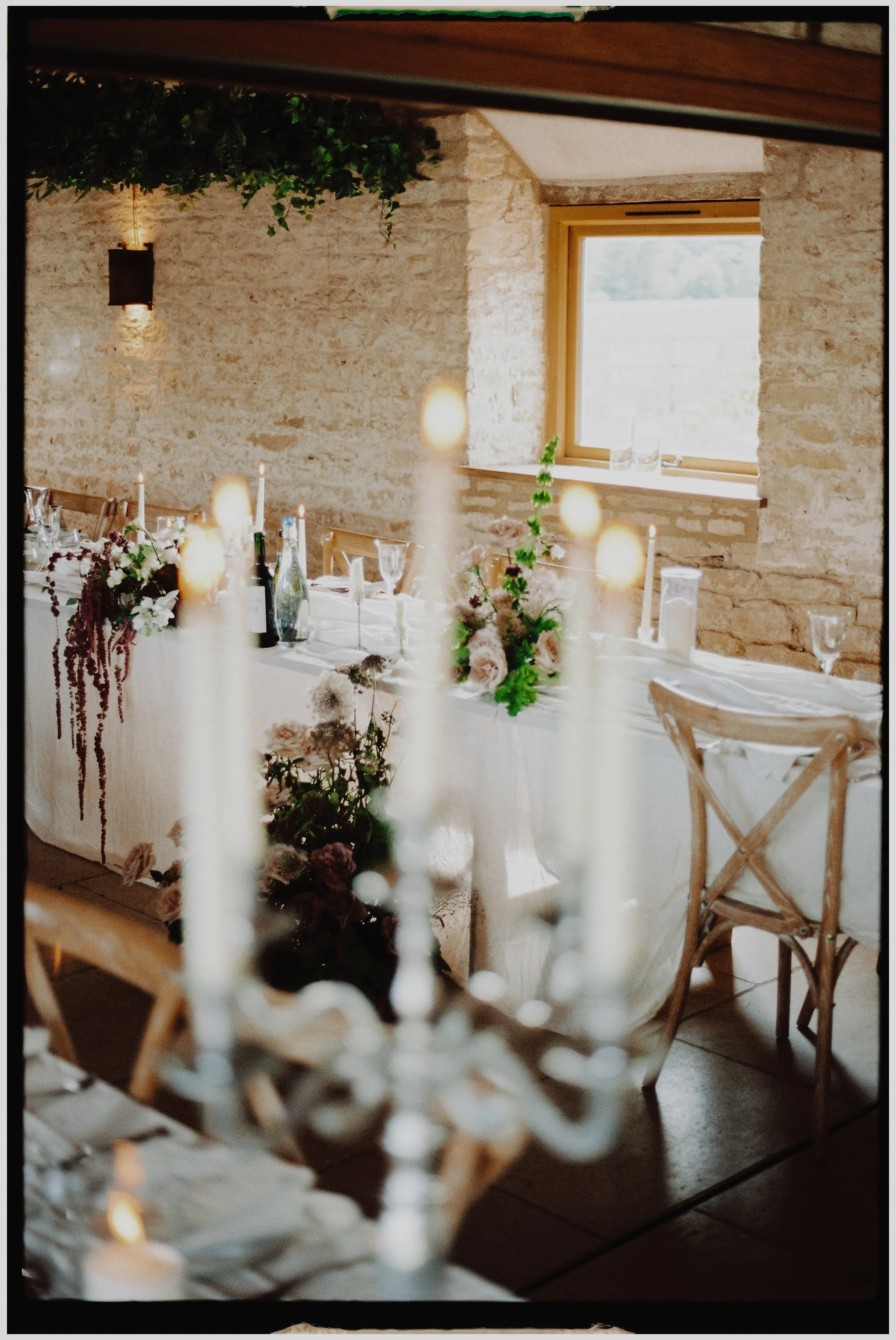 A decorated dining table with floral centerpieces, candles, wine bottles, and glassware in a rustic room with stone walls and a window.