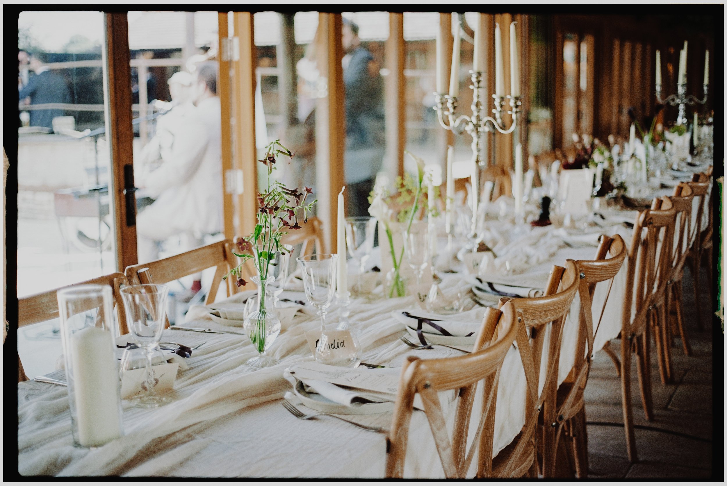 A long dining table set for a formal event with white tablecloths, decorated with candle holders, flowers, and glassware, near large windows with a view outside.