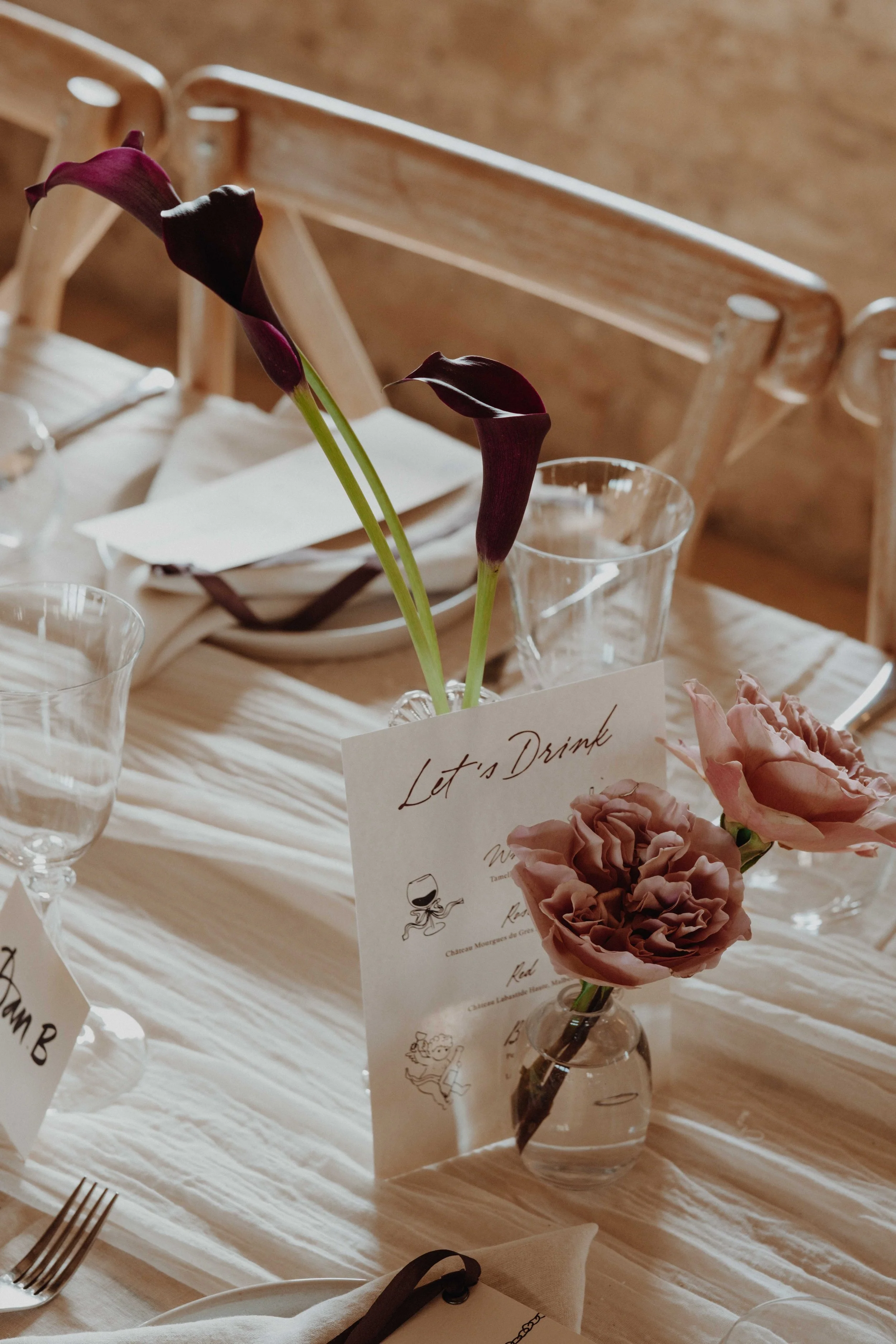 Table setting with a small vase of pink flowers, dark purple calla lilies, a handwritten card that says "Let's Dine," and empty glasses.
