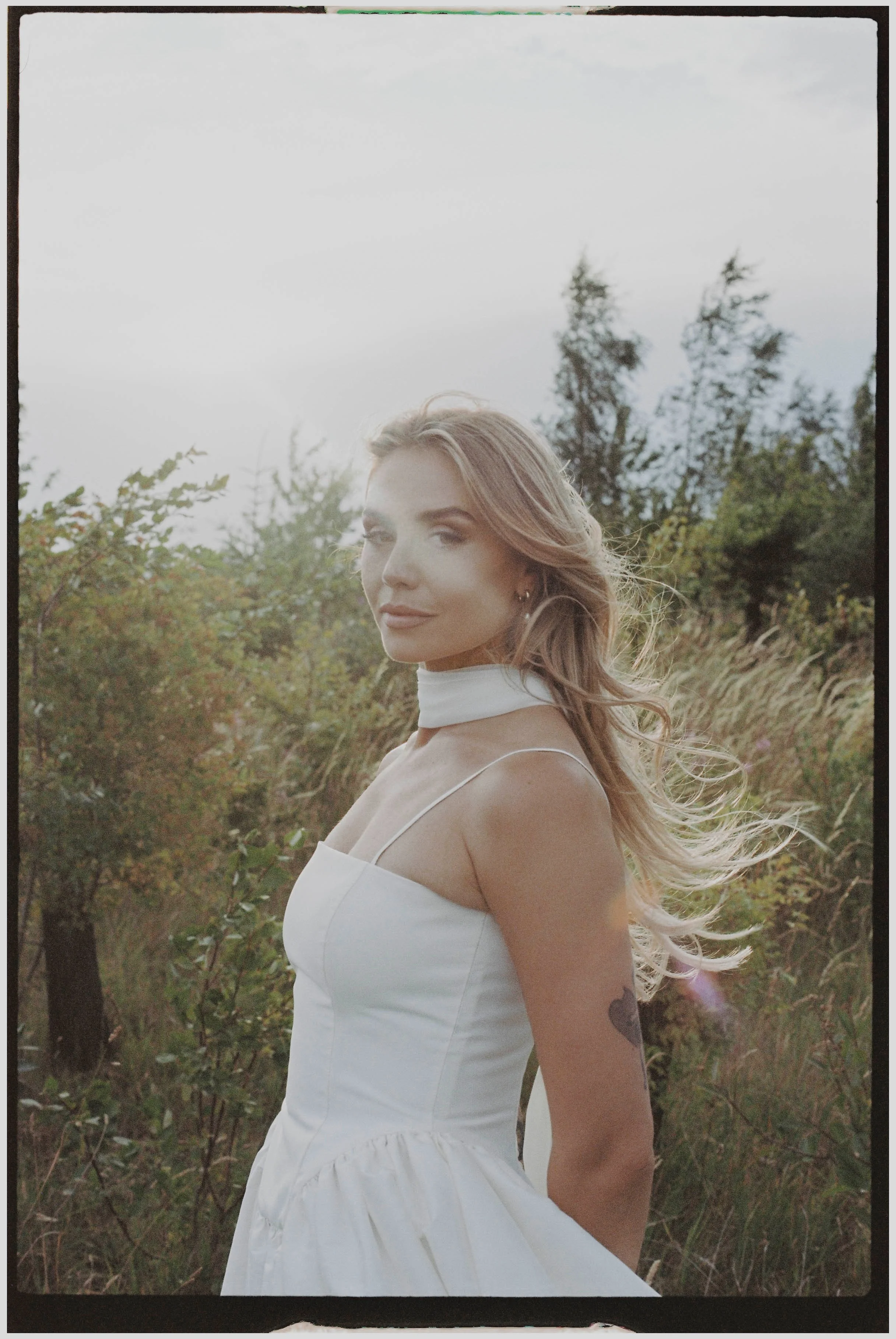 A woman with wavy light brown hair wearing a white dress outdoors in a field with tall grass and trees under a cloudy sky.