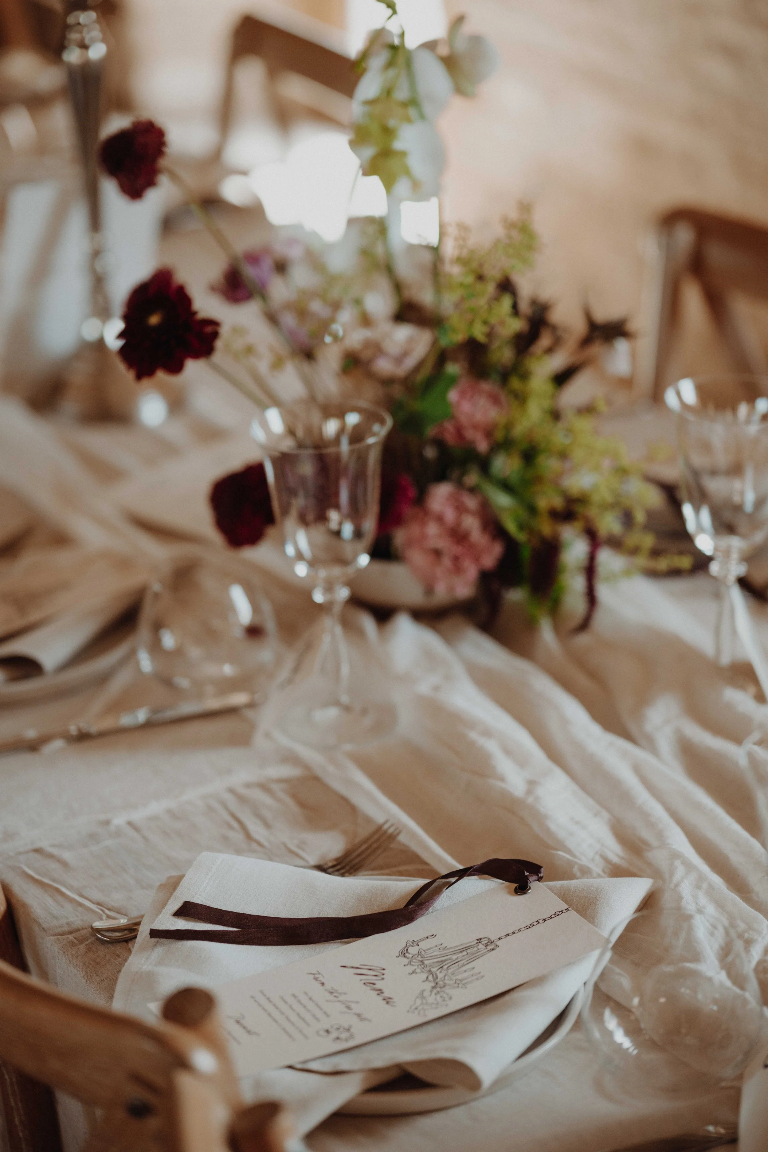 A table setting with a menu, glassware, and a floral centerpiece on a beige tablecloth.