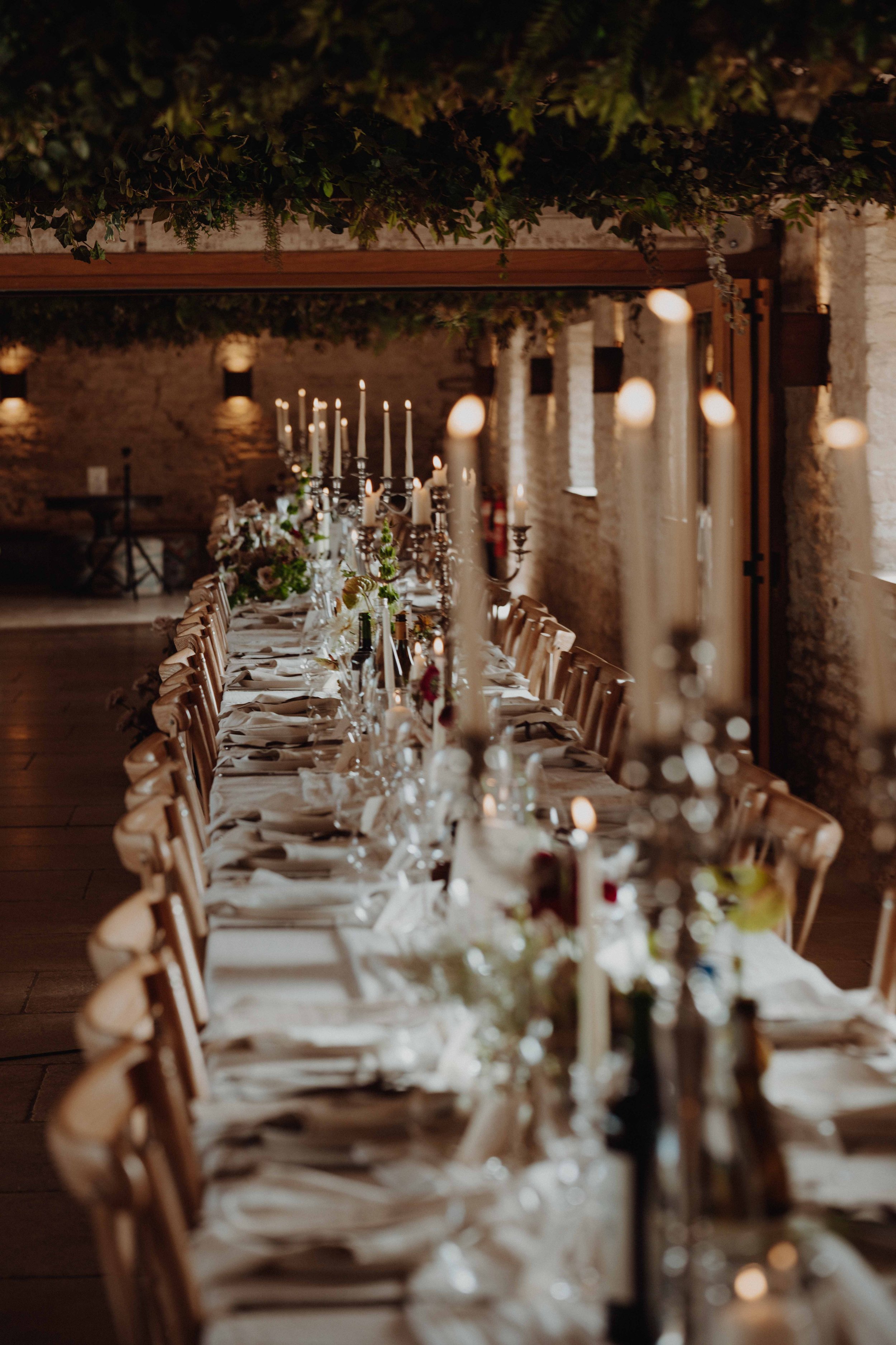 Elegantly decorated long dinner table set with white tablecloths, candles, and floral centerpieces in a rustic stone-walled reception hall.