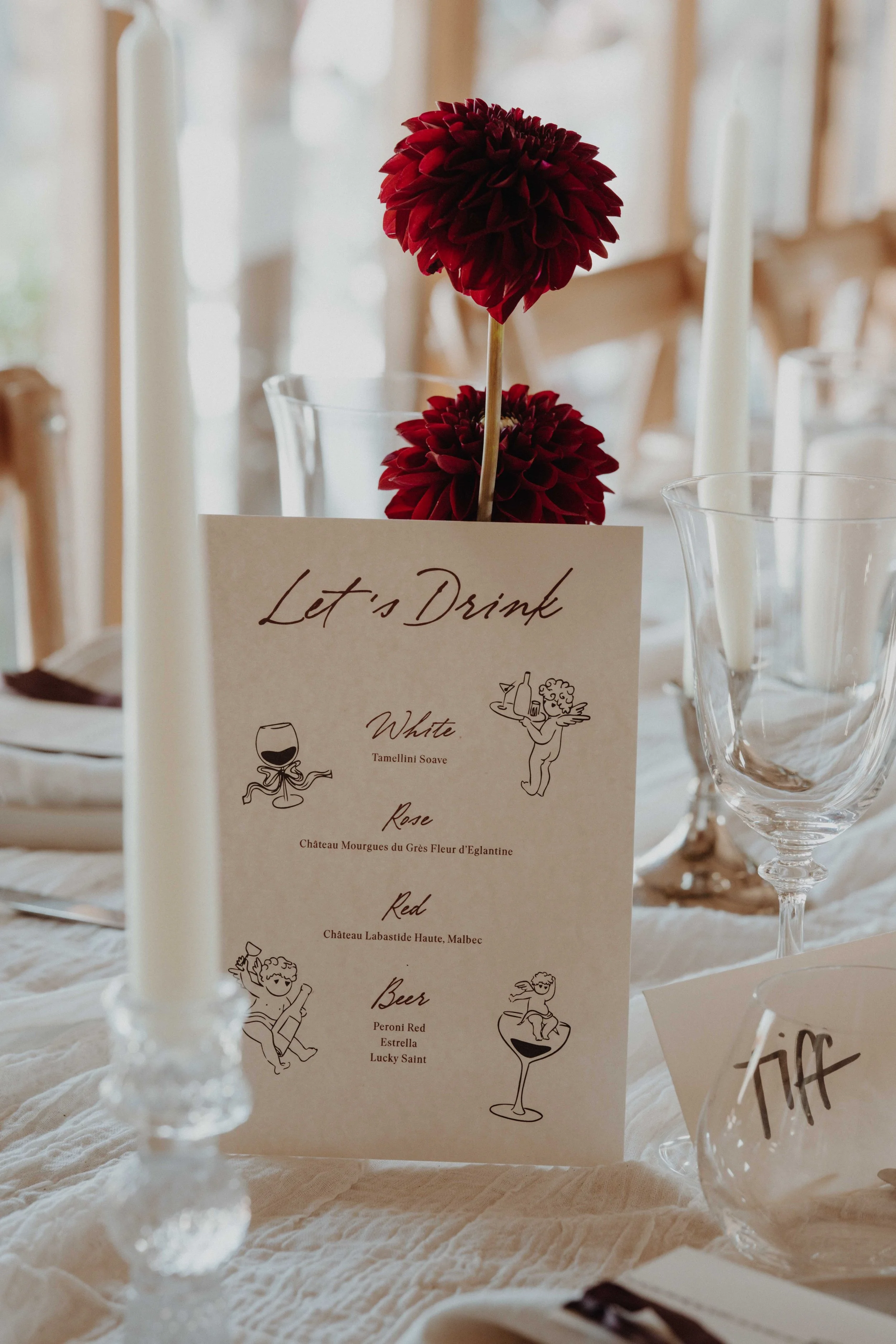 A dinner table setting with a menu card titled "Let's Drink" listing wine, rose, red wine, and beer options, surrounded by candles, wine glasses, and a red flower centerpiece.