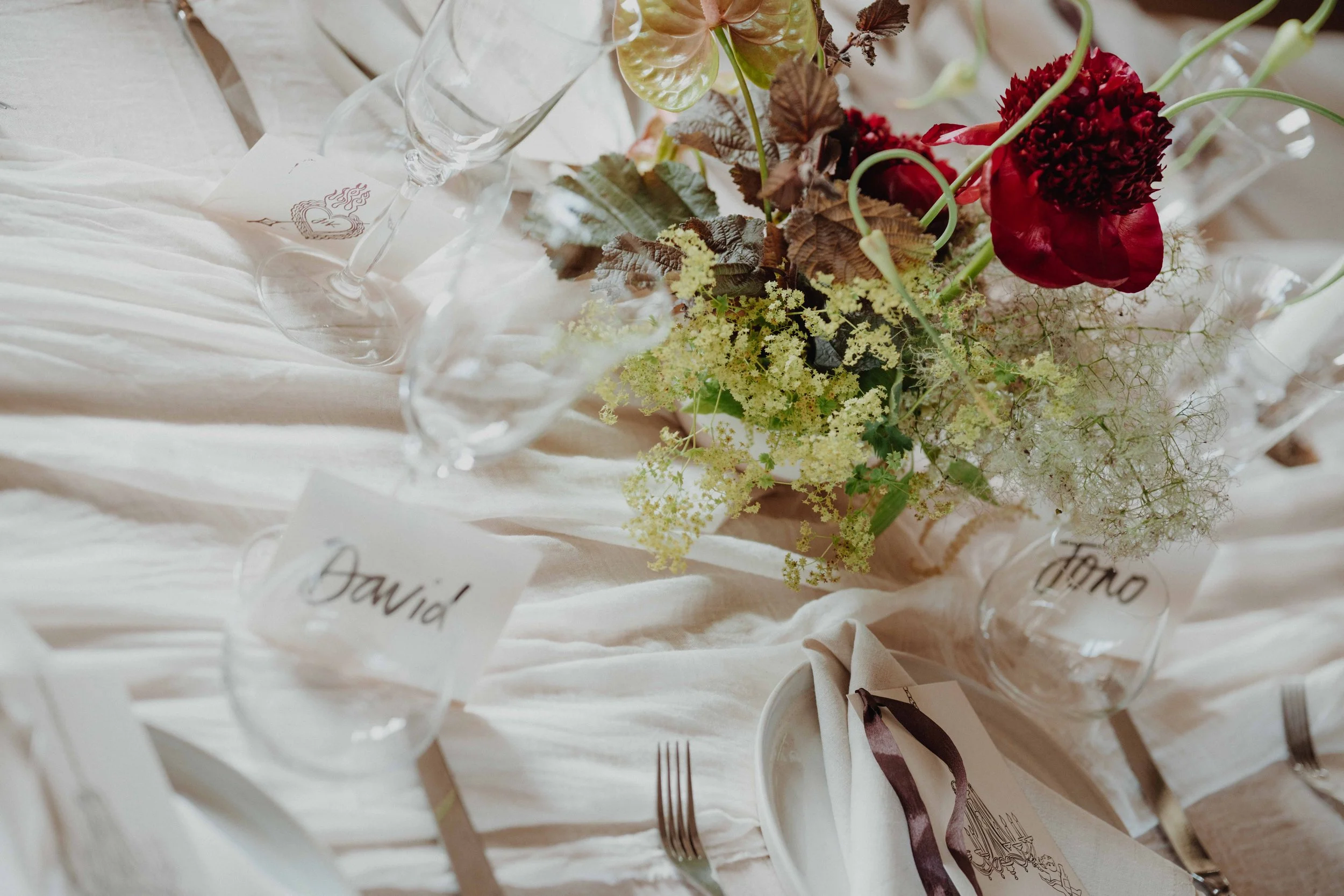 A table centerpiece with a lush floral arrangement of red and yellow flowers and greenery, set on a white tablecloth with glassware, a name card labeled 'David', and a napkin with a dark ribbon.