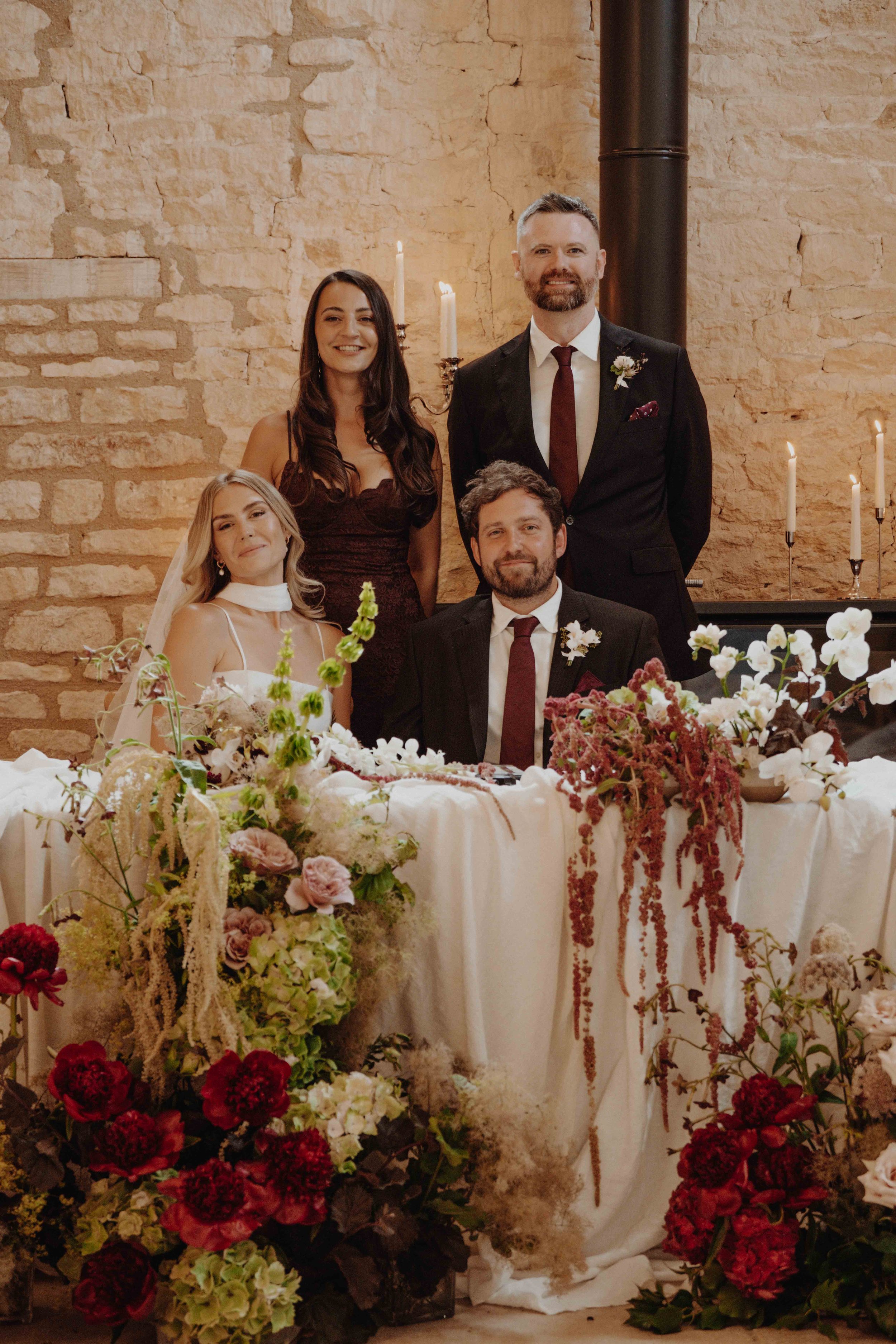 A wedding celebration with four people posing behind a decorated table. The bride is seated on the left, wearing a white wedding dress and veil. The groom is seated next to her, wearing a black suit with a white shirt, burgundy tie, and boutonniere. 
