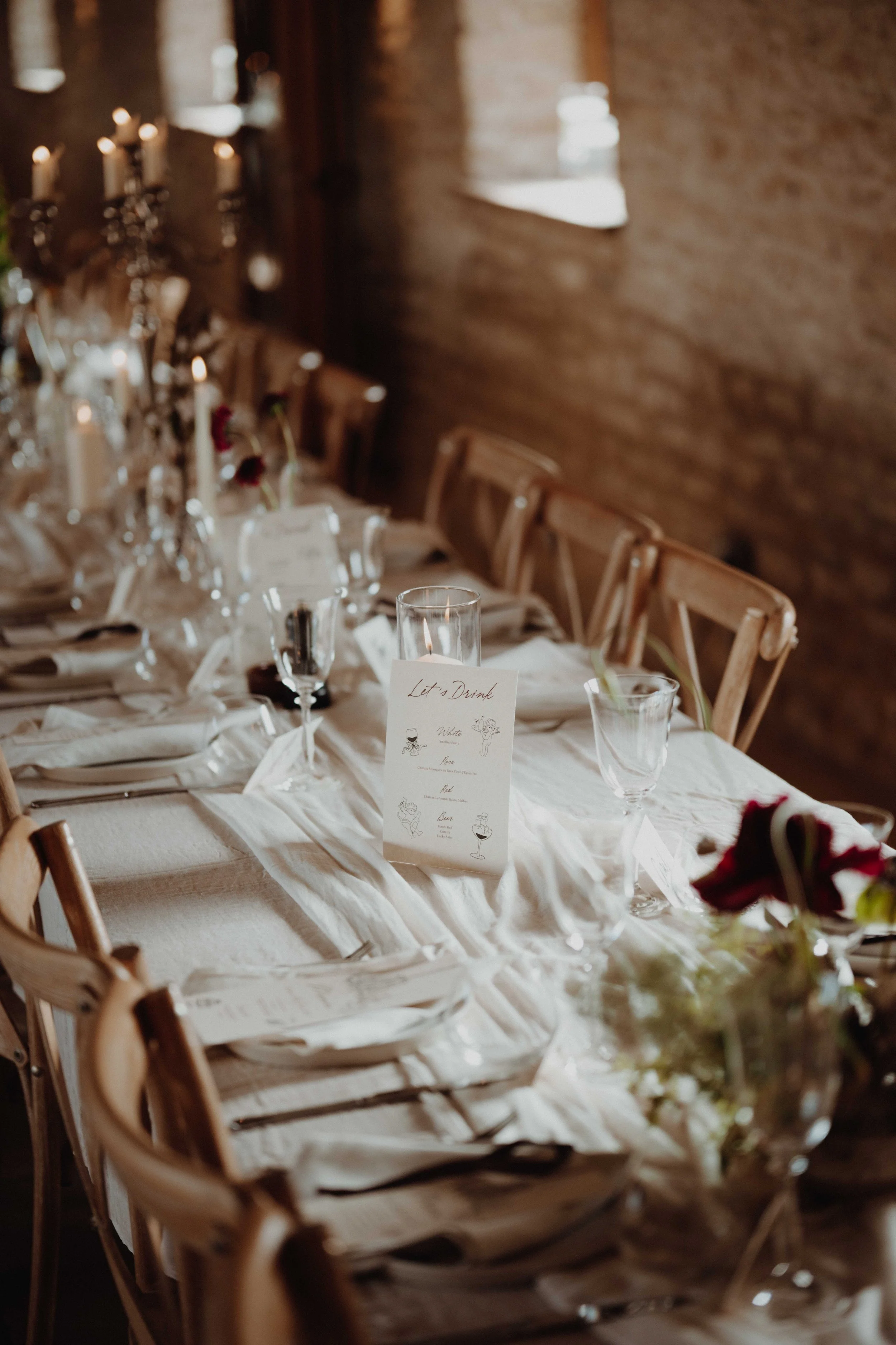 Elegant dining table set for a celebration or wedding, decorated with candles, flower arrangements, and a 'Let's Drink' menu card in a rustic room with brick walls.