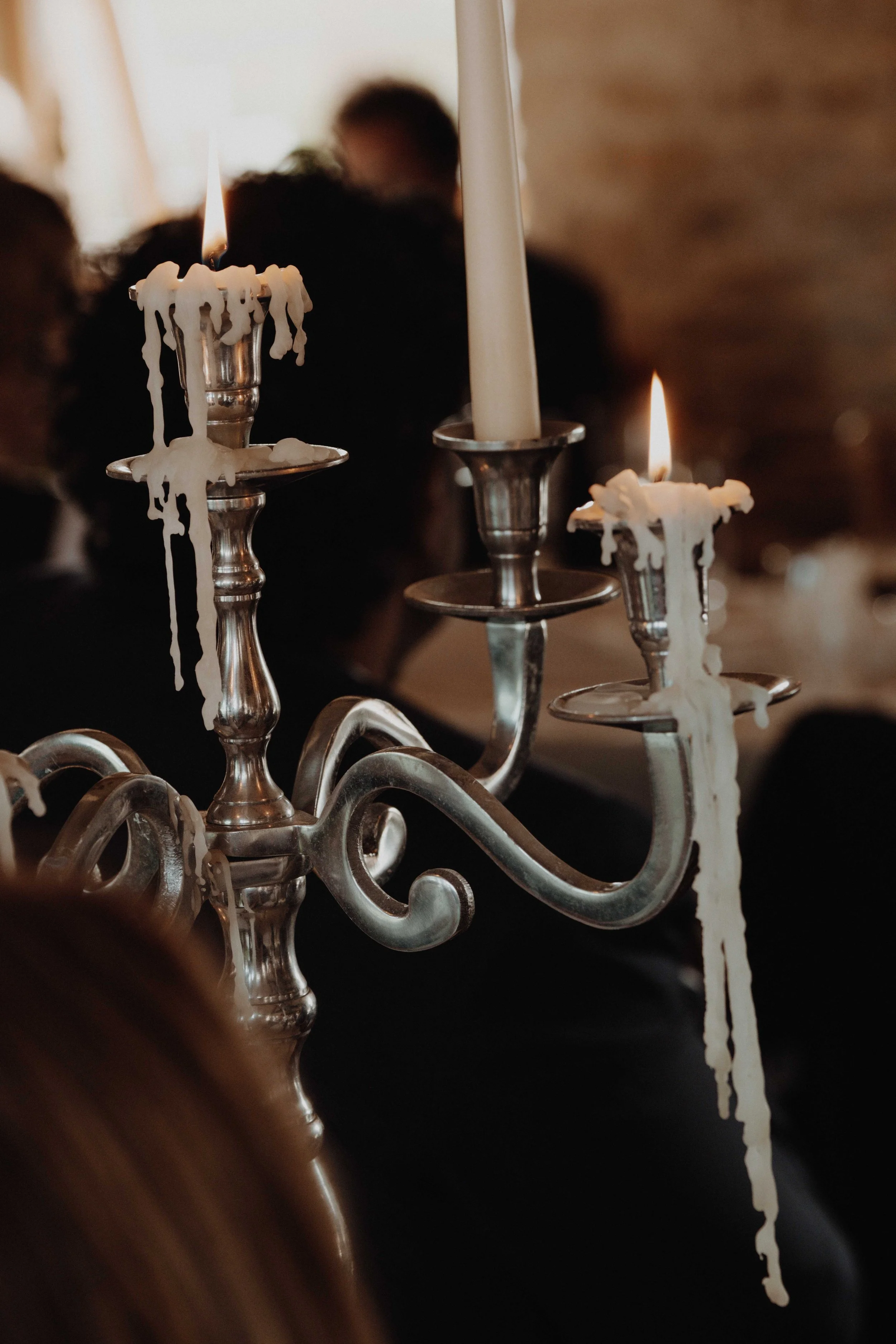 A silver candelabrum with three lit white candles, some of which are melting and dripping wax. The background shows blurry guests in a dimly lit setting.