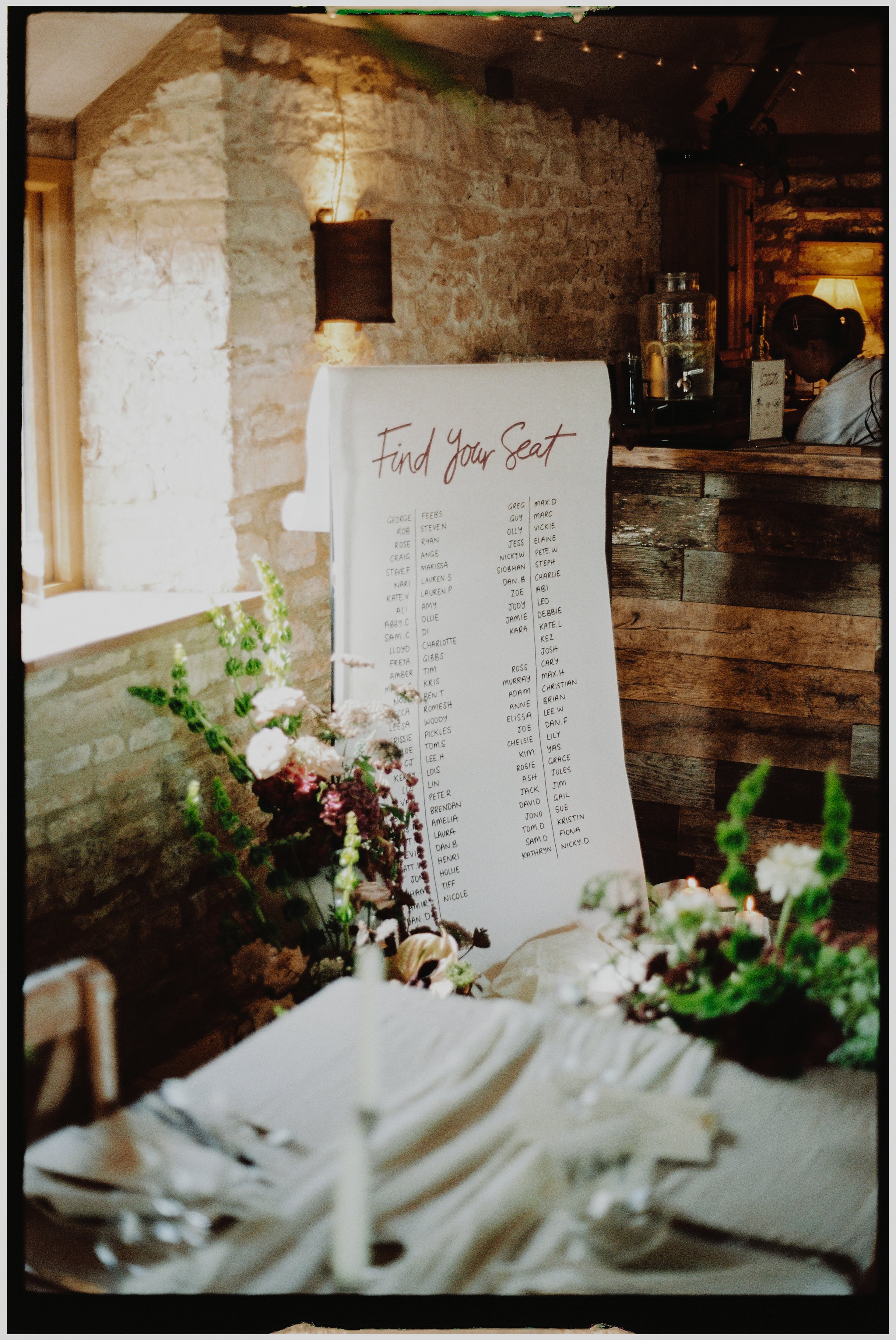 Wedding reception seating chart with the words 'Find Your Seat' at the top, displayed on a white board in a rustic setting with stone and wood walls, and decorated with flowers and candles.