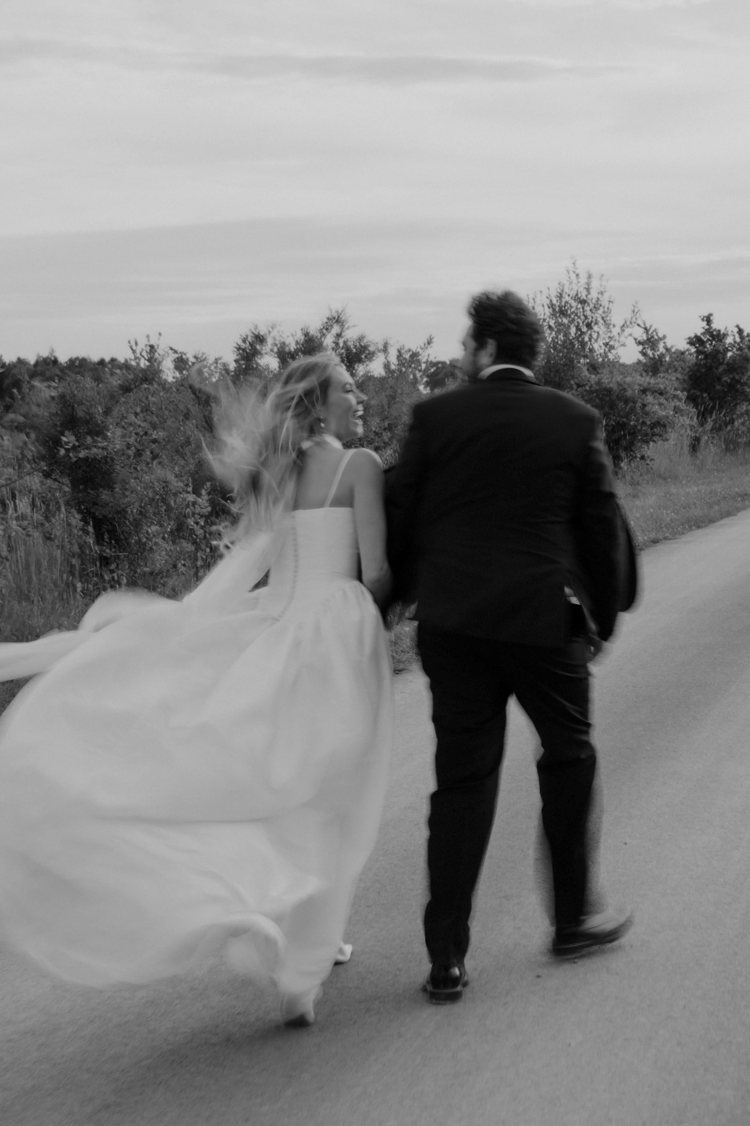A bride and groom running down a road, holding hands, with trees and sky in the background, in black and white.