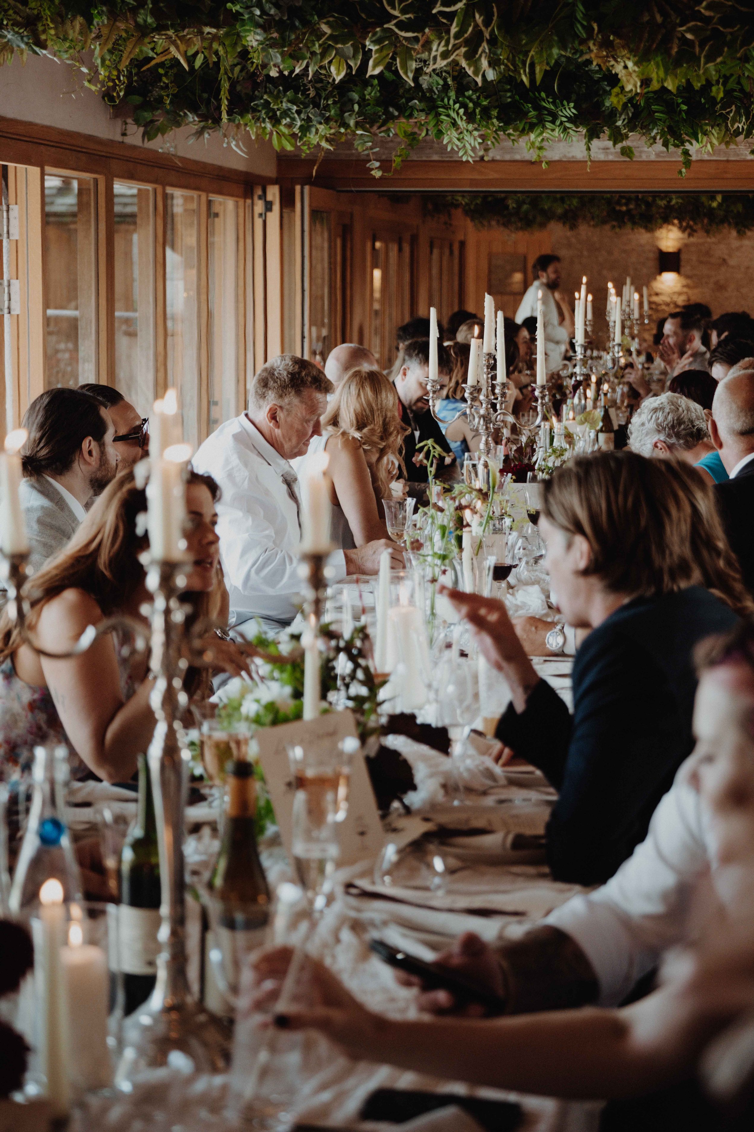 A large group of people sitting around a long dining table at a formal event or wedding reception in a warmly lit, rustic indoor setting with wooden walls and greenery ceiling decor.