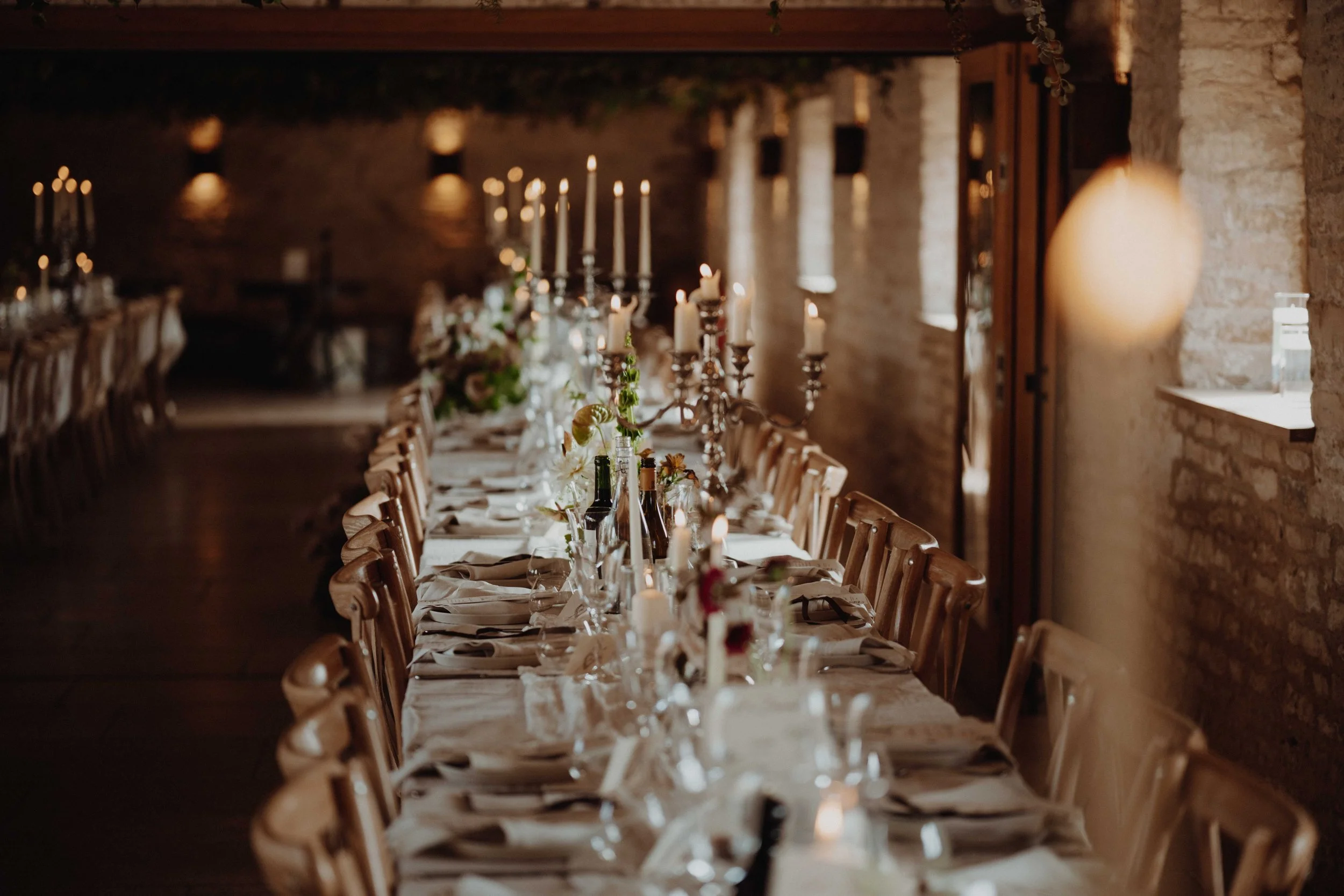 Long dining table decorated with candles, flowers, and glassware, set for a formal event in a rustic, brick-walled room with warm lighting.