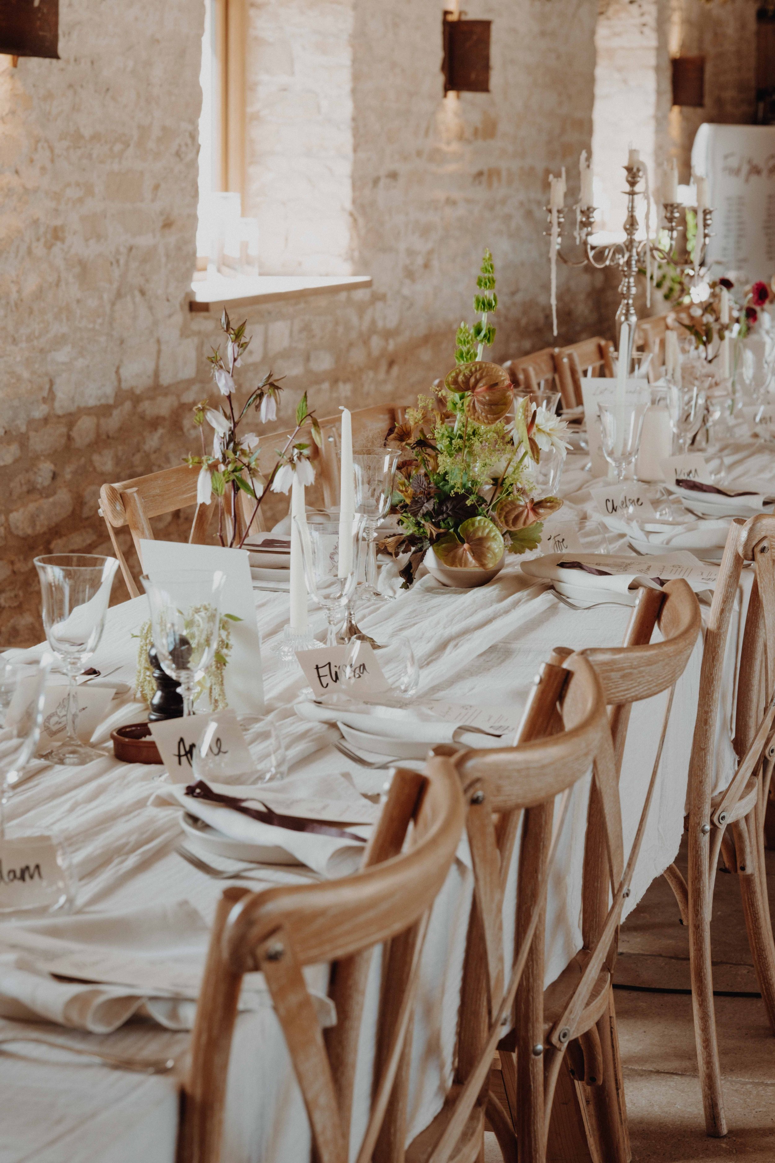 A rustic dining table set for a formal event with floral centerpiece, candles, place cards, and white tablecloth, against a brick wall.