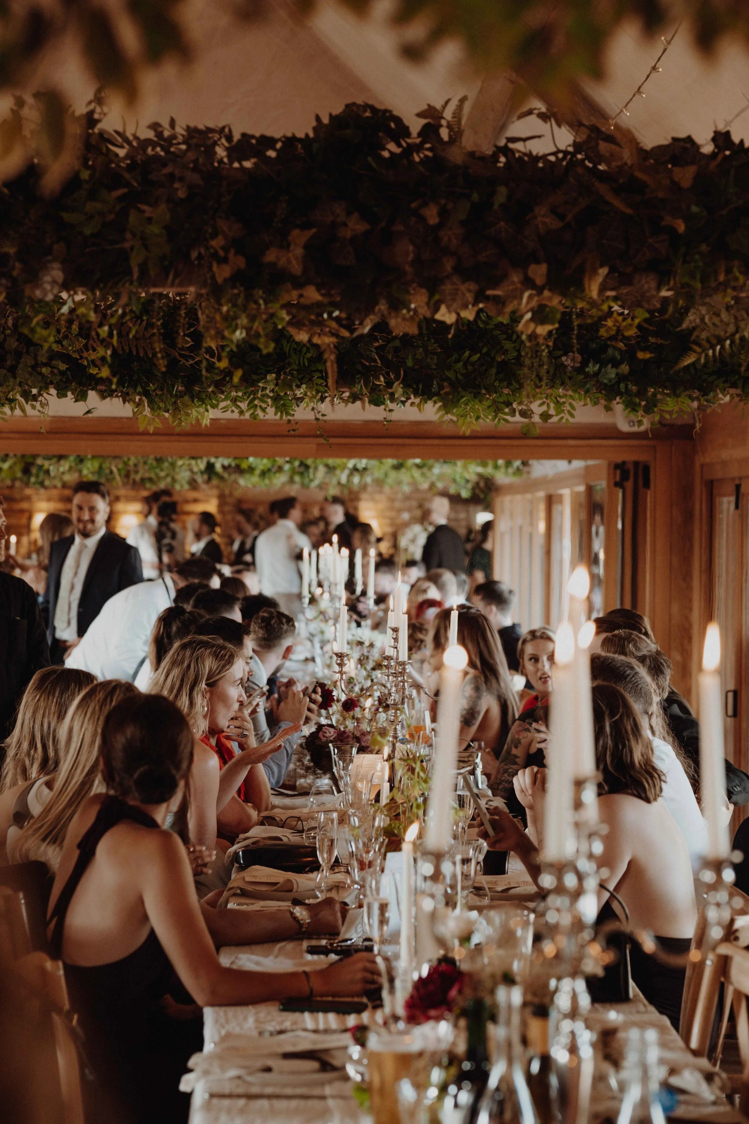 A long dinner table set for a wedding or celebration with candles, floral centerpieces, and guests seated and socializing inside a wood-paneled room decorated with greenery on the ceiling.