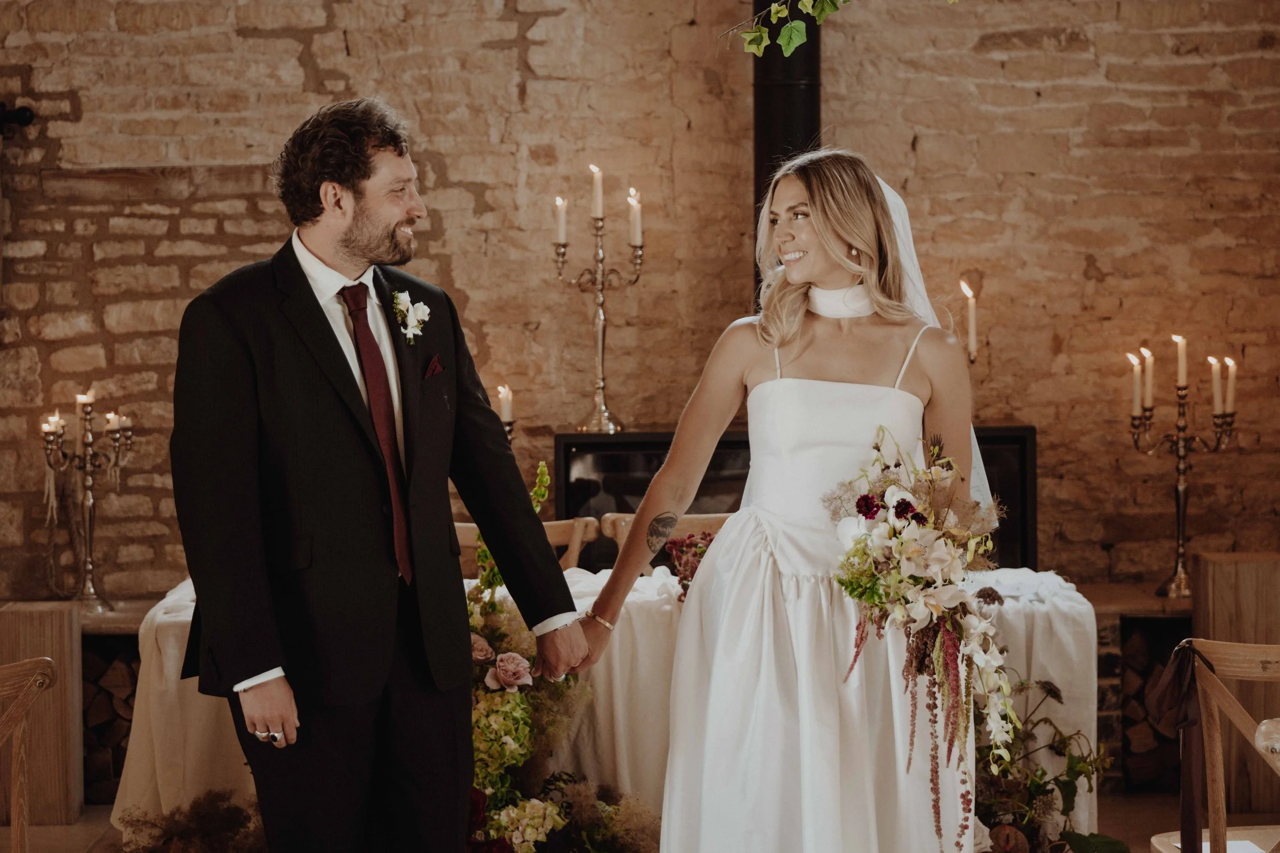 A bride and groom holding hands and smiling at each other during their wedding ceremony inside a rustic venue with exposed brick walls and candlelit chandeliers.