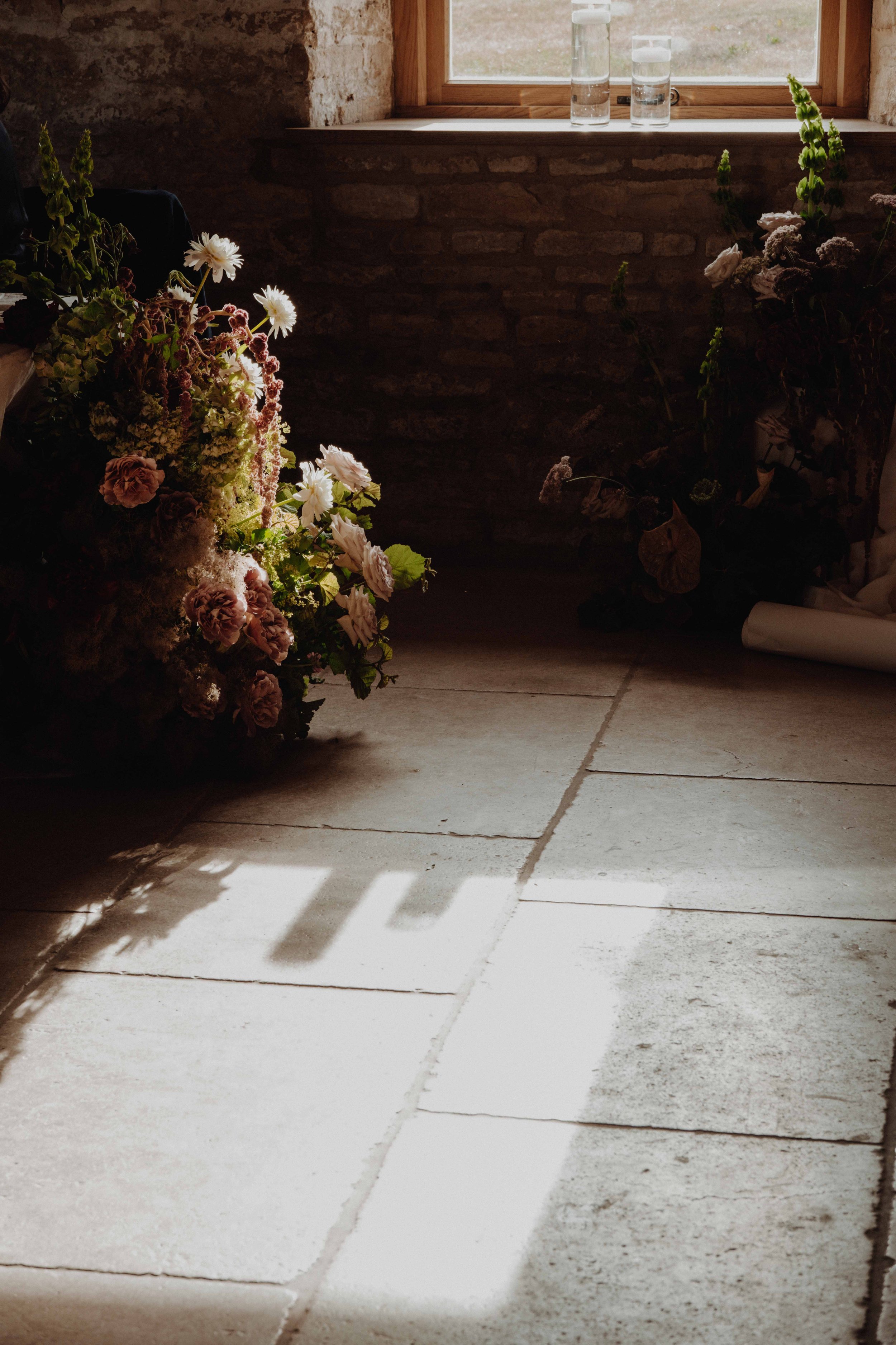 Sunlight casting over a white tiled floor with floral arrangements near a brick wall and window.