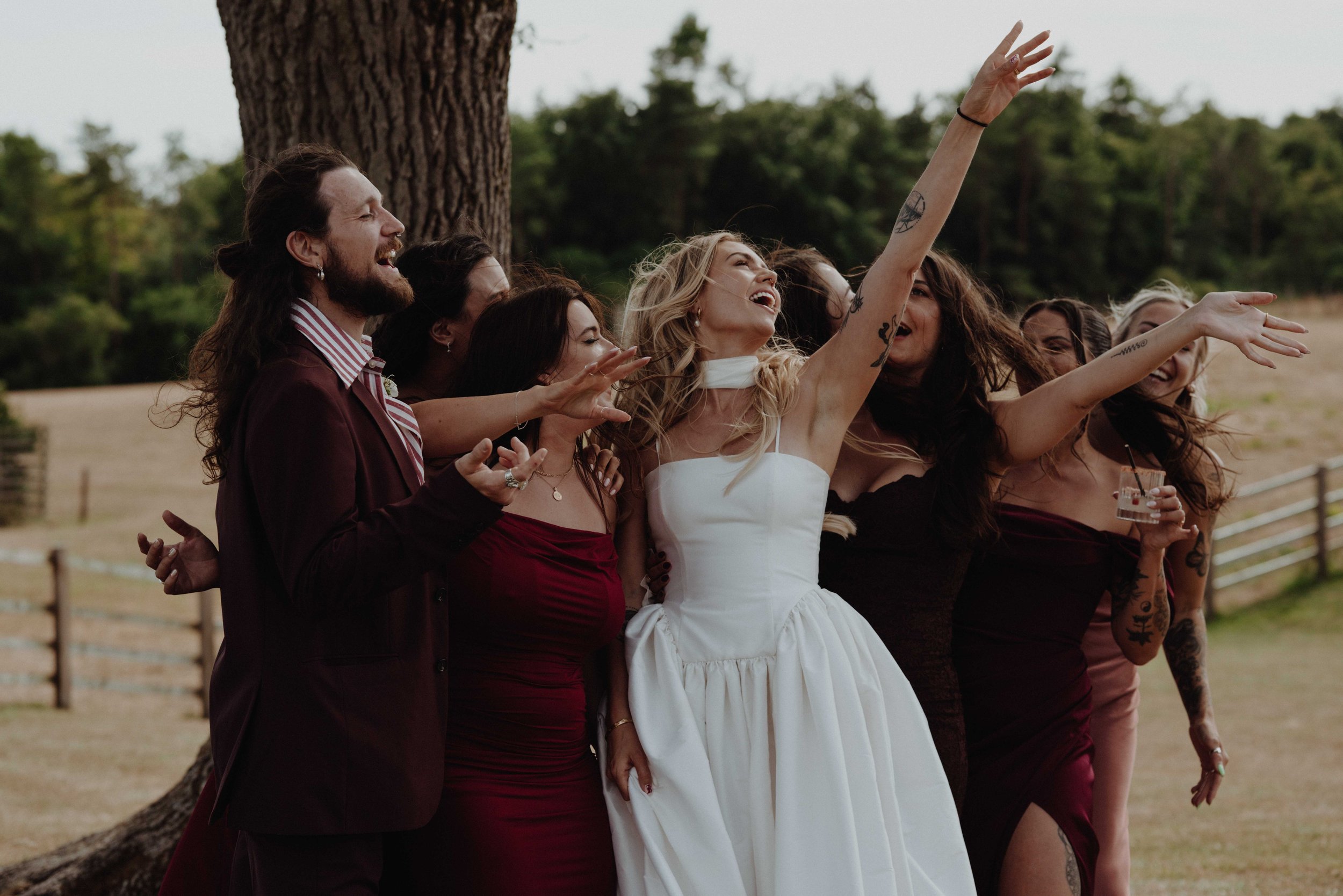 Group of people celebrating outdoors, with a woman in a white dress reaching up and a man in a suit smiling, surrounded by friends or family.