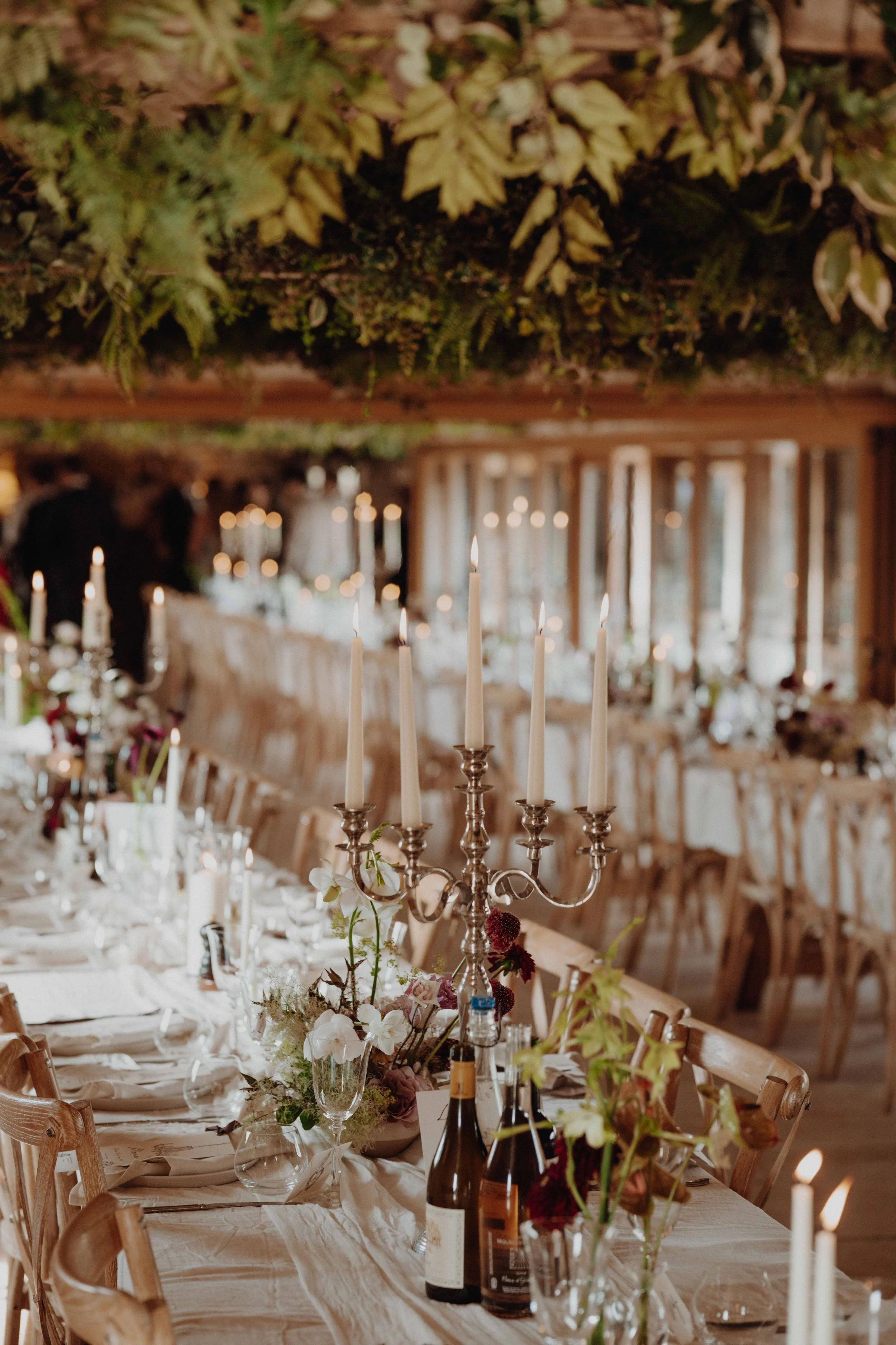 Elegant dining table set with candles, flowers, and wine bottles, decorated for a special event in a warmly lit room with windows.