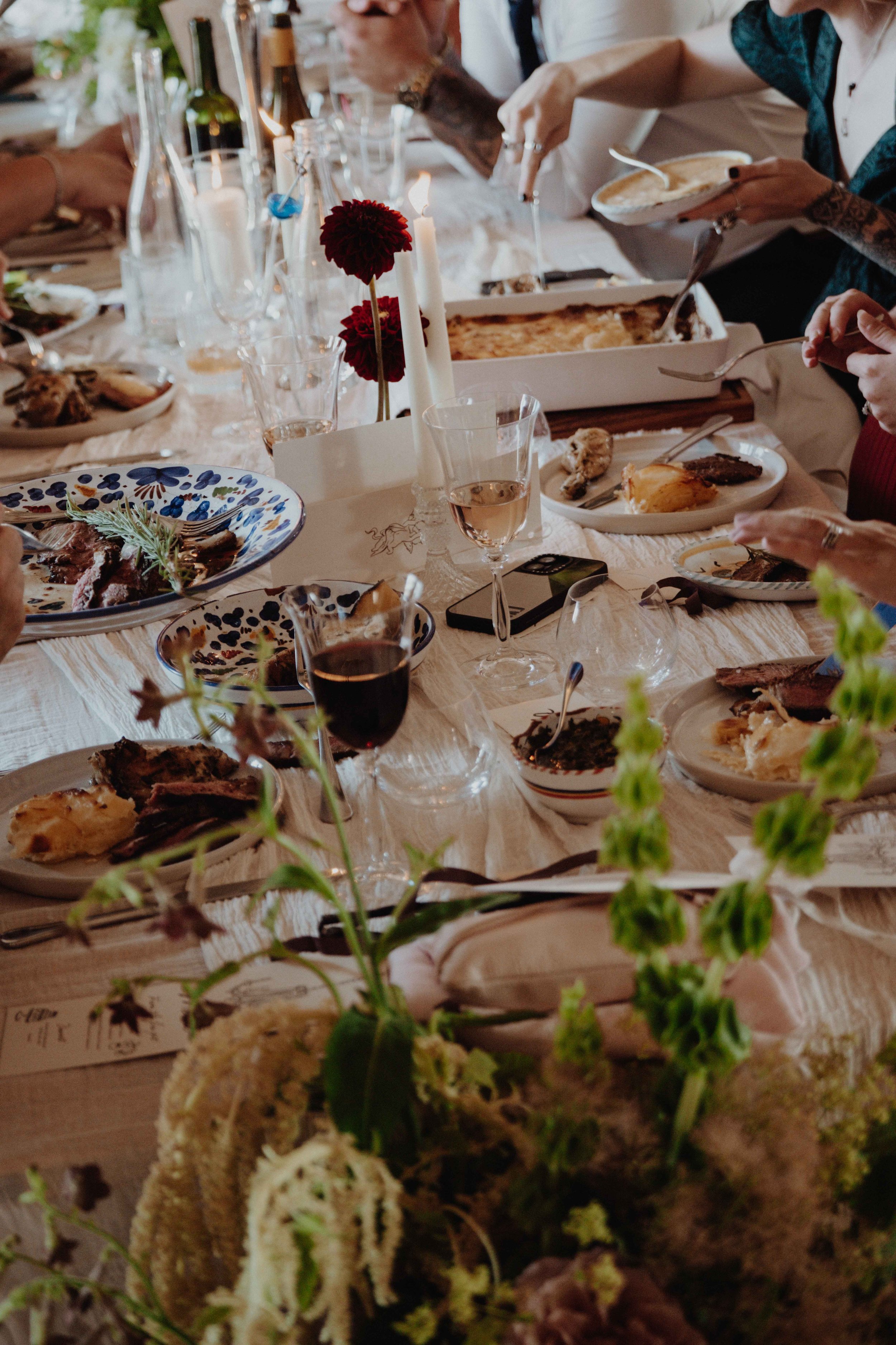 A family gathering around a dining table with plates of food, wine glasses, and a centerpiece with red flowers and candles.