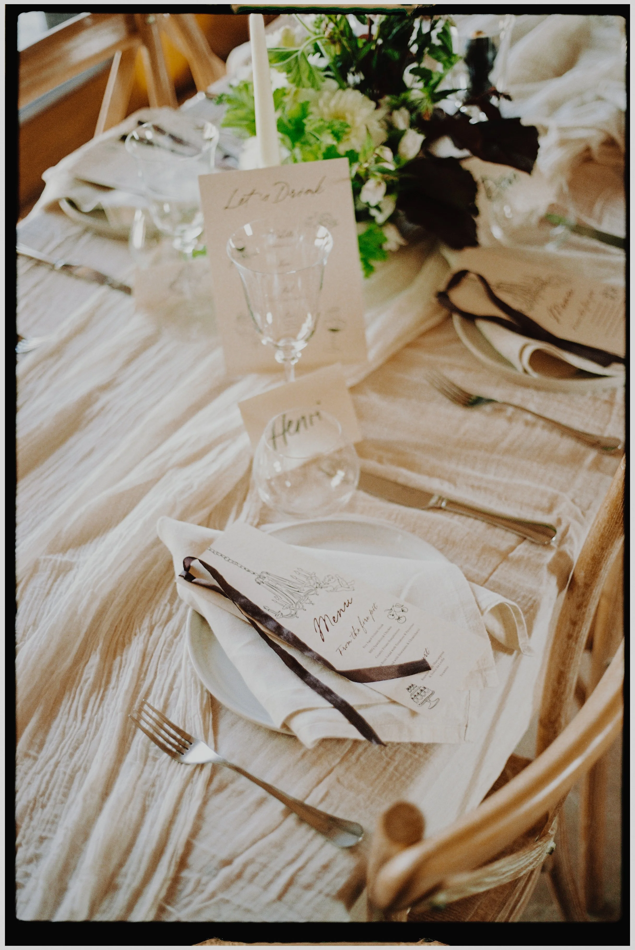 A rustic wedding table setting with a cream-colored tablecloth, glassware, a menu card, a place card, silverware, a napkin, and a floral centerpiece with green and white flowers.