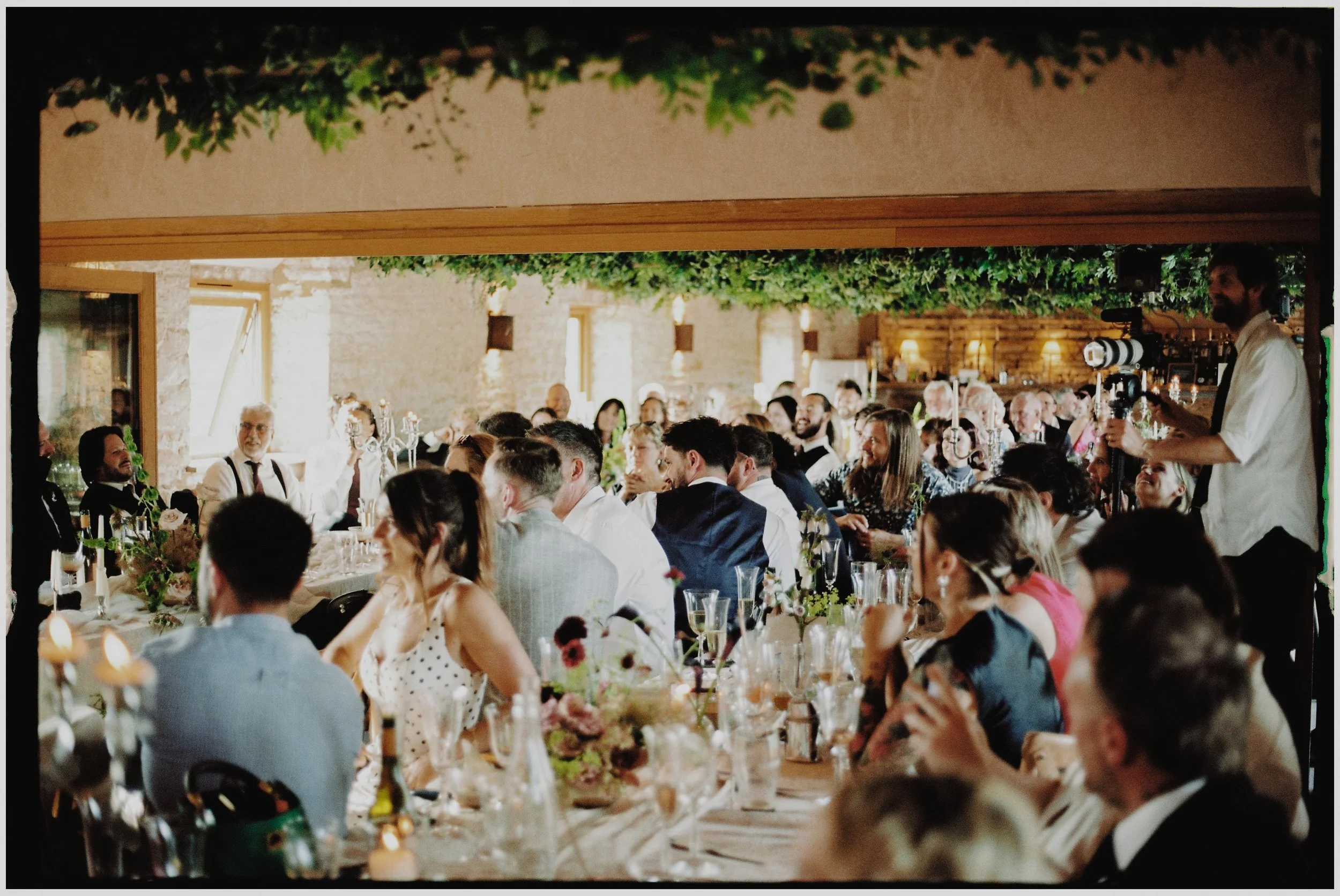 People attending a wedding reception or formal dinner in a rustic indoor venue with stone walls, greenery ceiling decor, and wooden accents, seated at long tables with floral centerpieces and glassware.