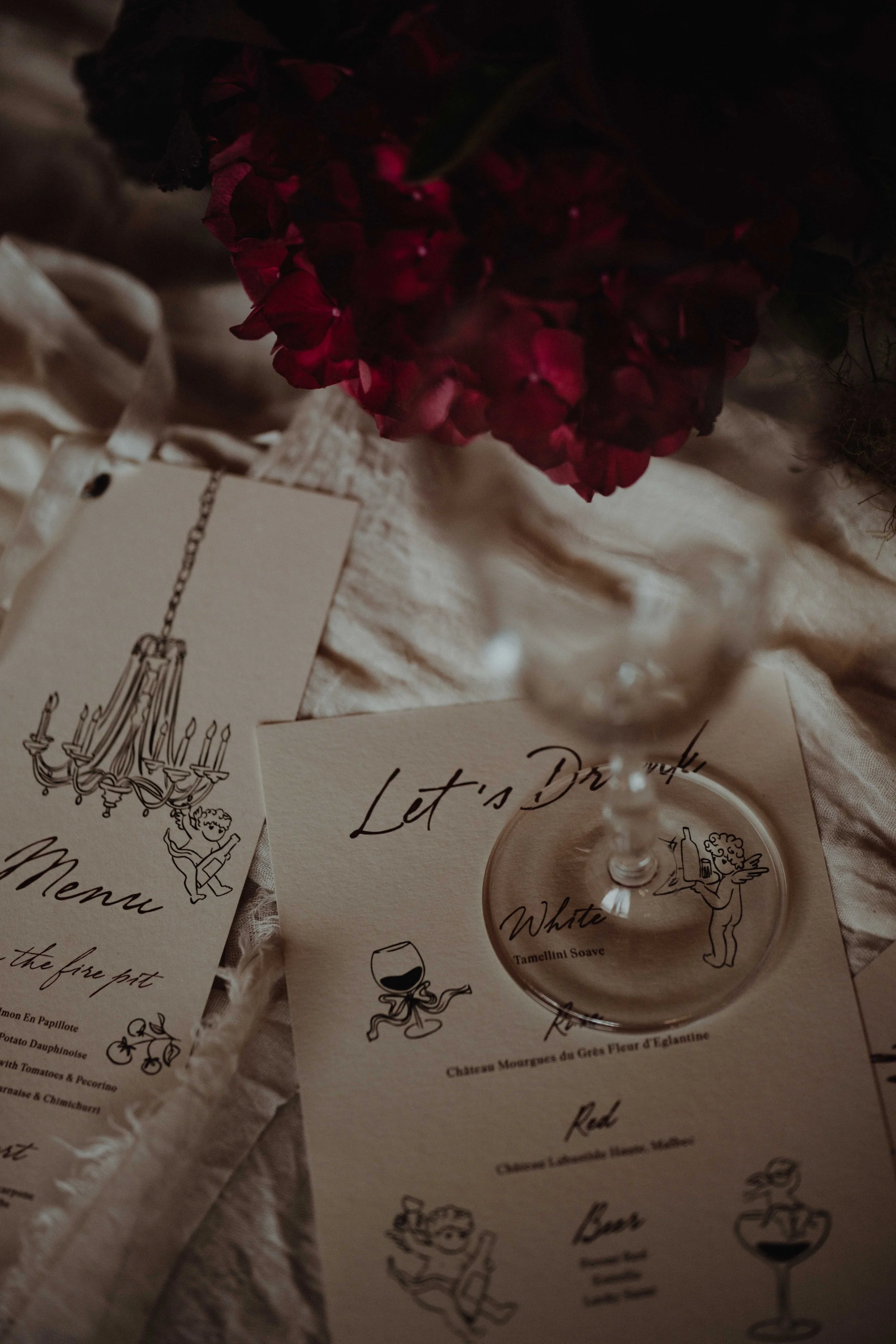 A dining scene with a menu, cocktail list, a wine glass, and a flower arrangement on a crumpled white tablecloth.