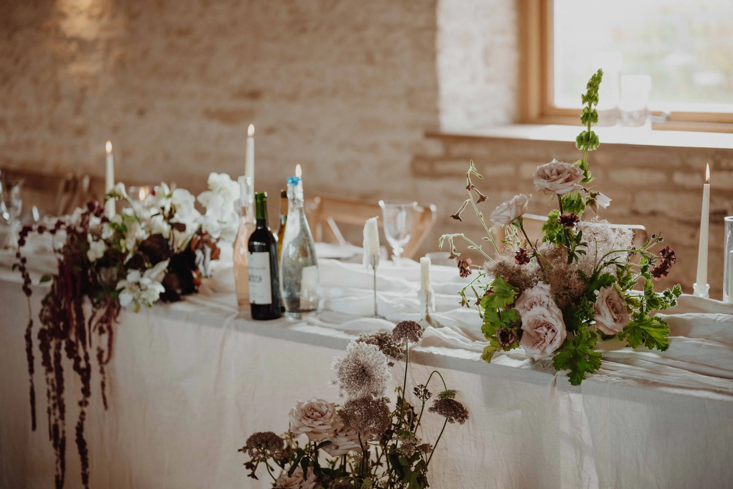 Decorated banquet table with candles, wine bottles, and floral arrangements in a rustic room with exposed brick wall