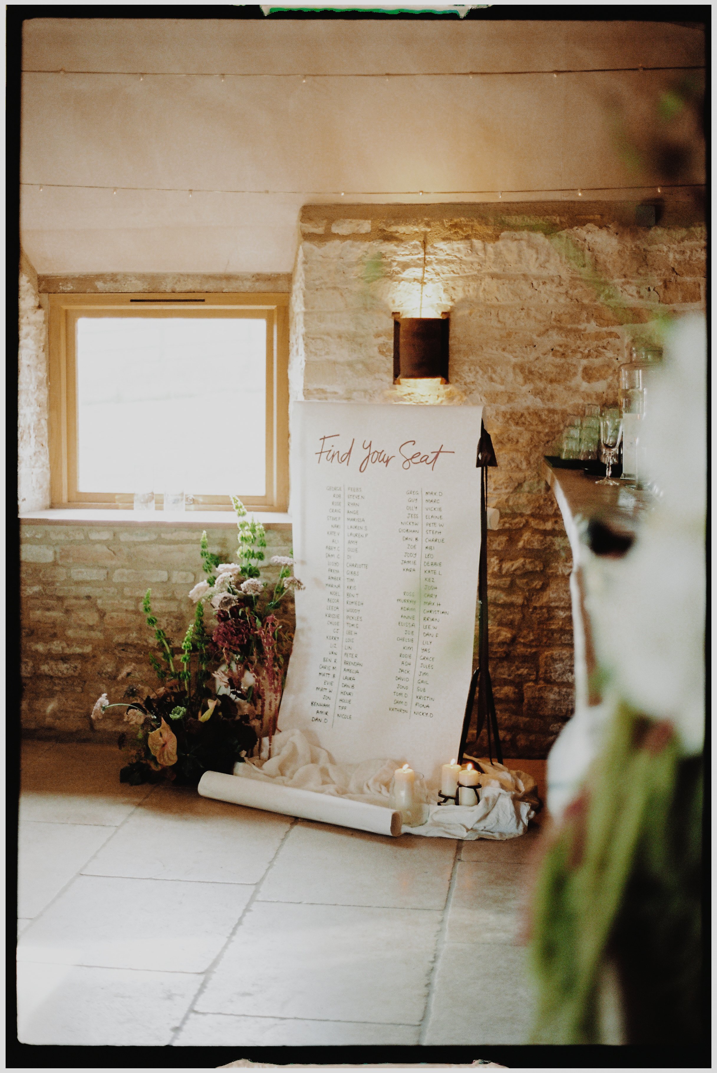 Wedding seating chart on a whiteboard with handwritten names under the heading 'Find Your Seat,' surrounded by candles, flowers, and glassware, in a rustic stone-walled venue with a window.