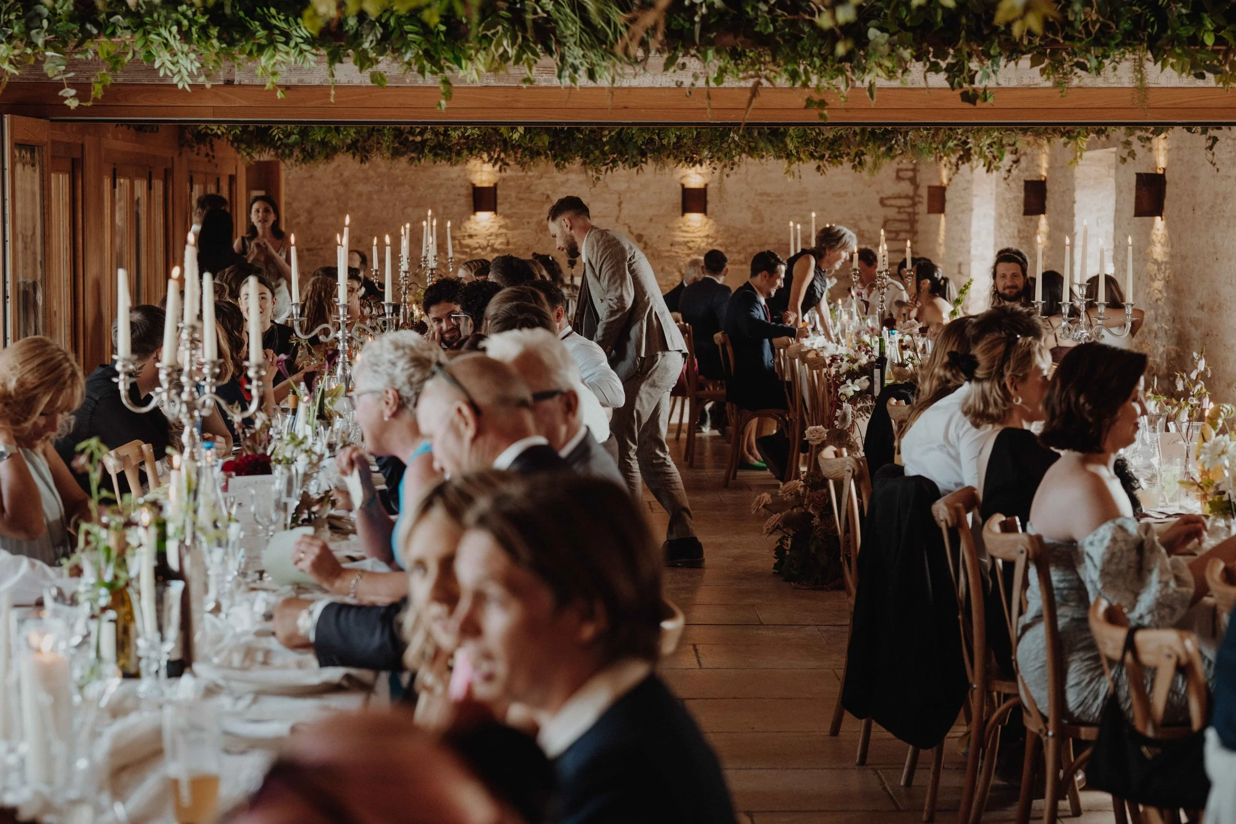 People seated at a long banquet table during a wedding reception in a rustic venue with candle centerpieces and floral decorations.