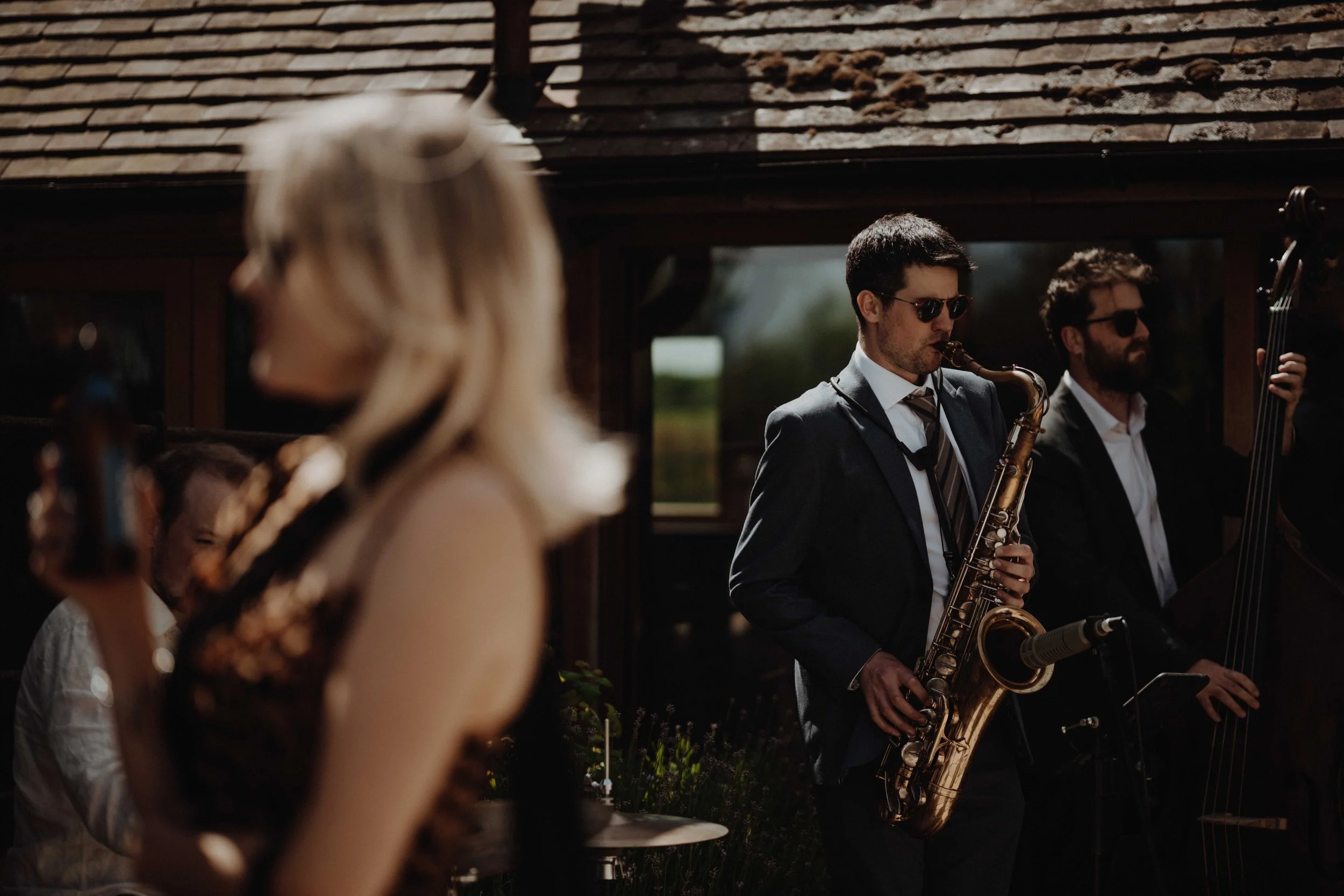 Musicians performing outdoors, with a woman in the foreground and two men playing saxophone and double bass in the background.